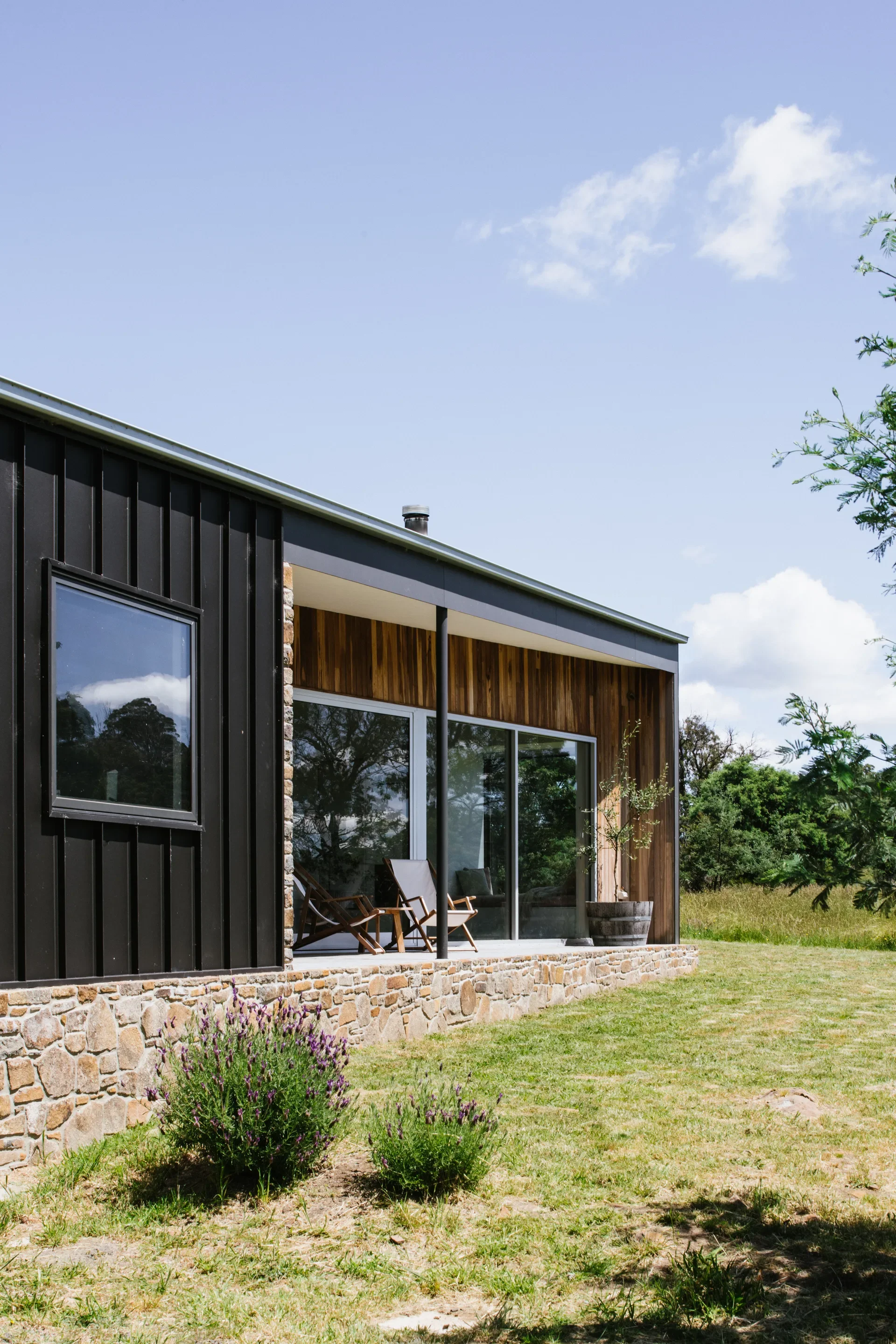 A dark, vertically-clad wall sits next to a timber-lined recessed patio with stone edging, where two wooden chairs overlook a sunny, grassy yard.