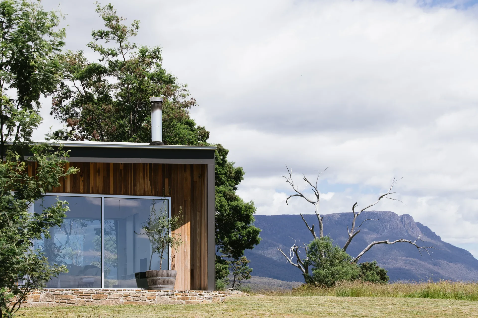 A timber-lined outdoor patio with a stone base features large glass doors and a silver chimney, looking out over a grassy field toward a distant mountain range.