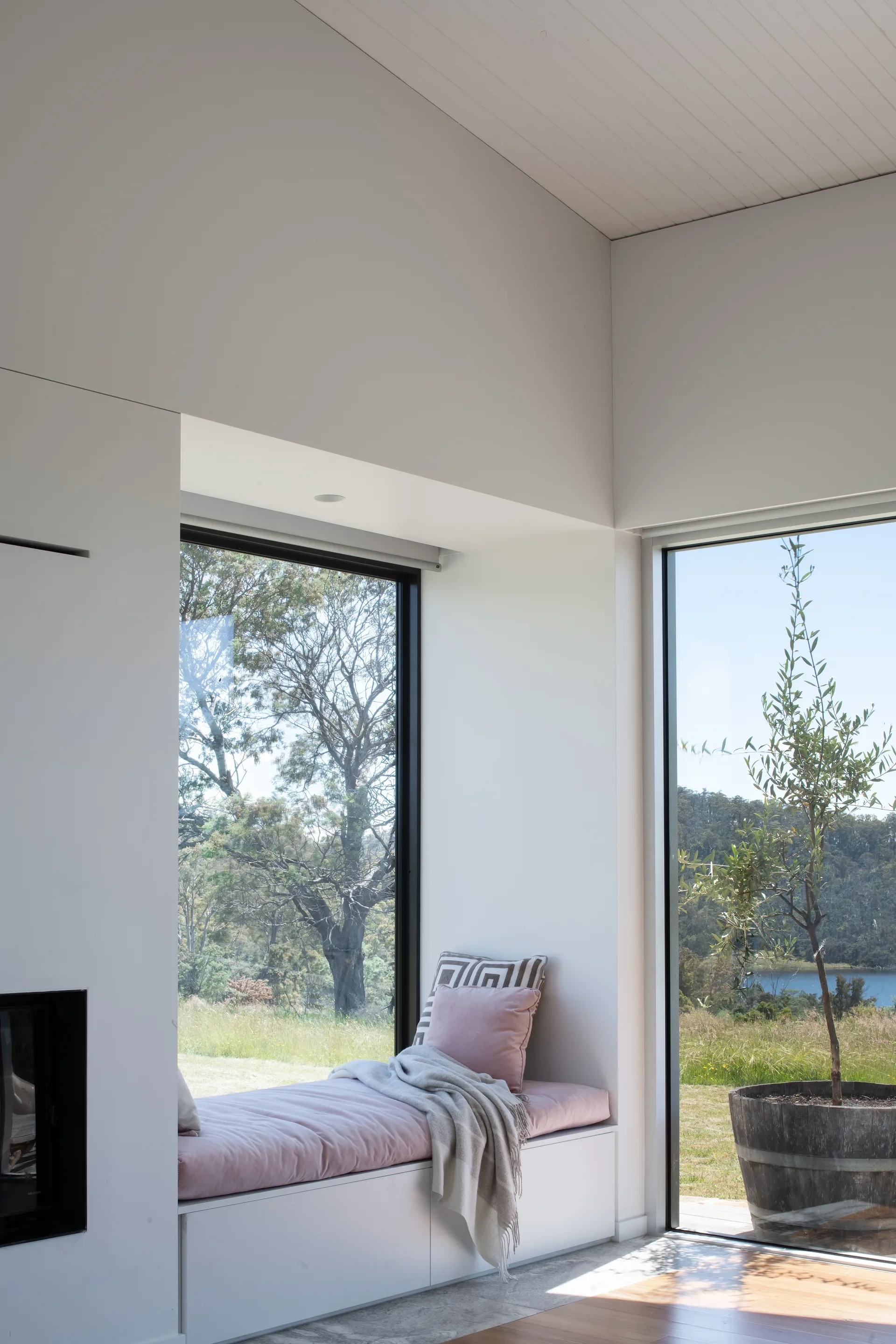 A recessed white window seat with a pink cushion and patterned pillow sits next to a large glass door, looking out at a potted olive tree and a distant lake.