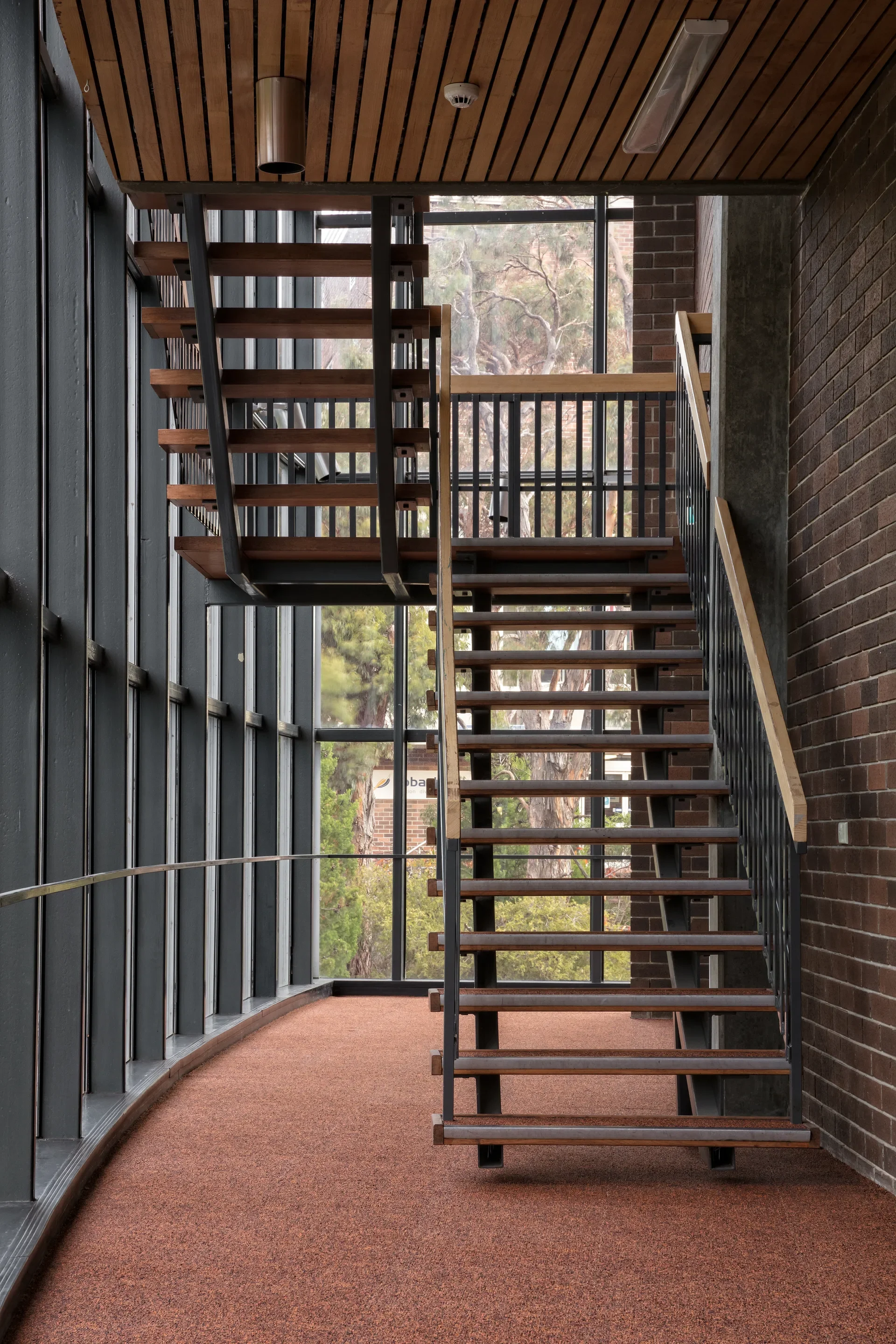 A wooden staircase with black metal stringers, on its right stands against a dark brick wall in a foyer with a reddish-brown textured floor and large windows to its left.