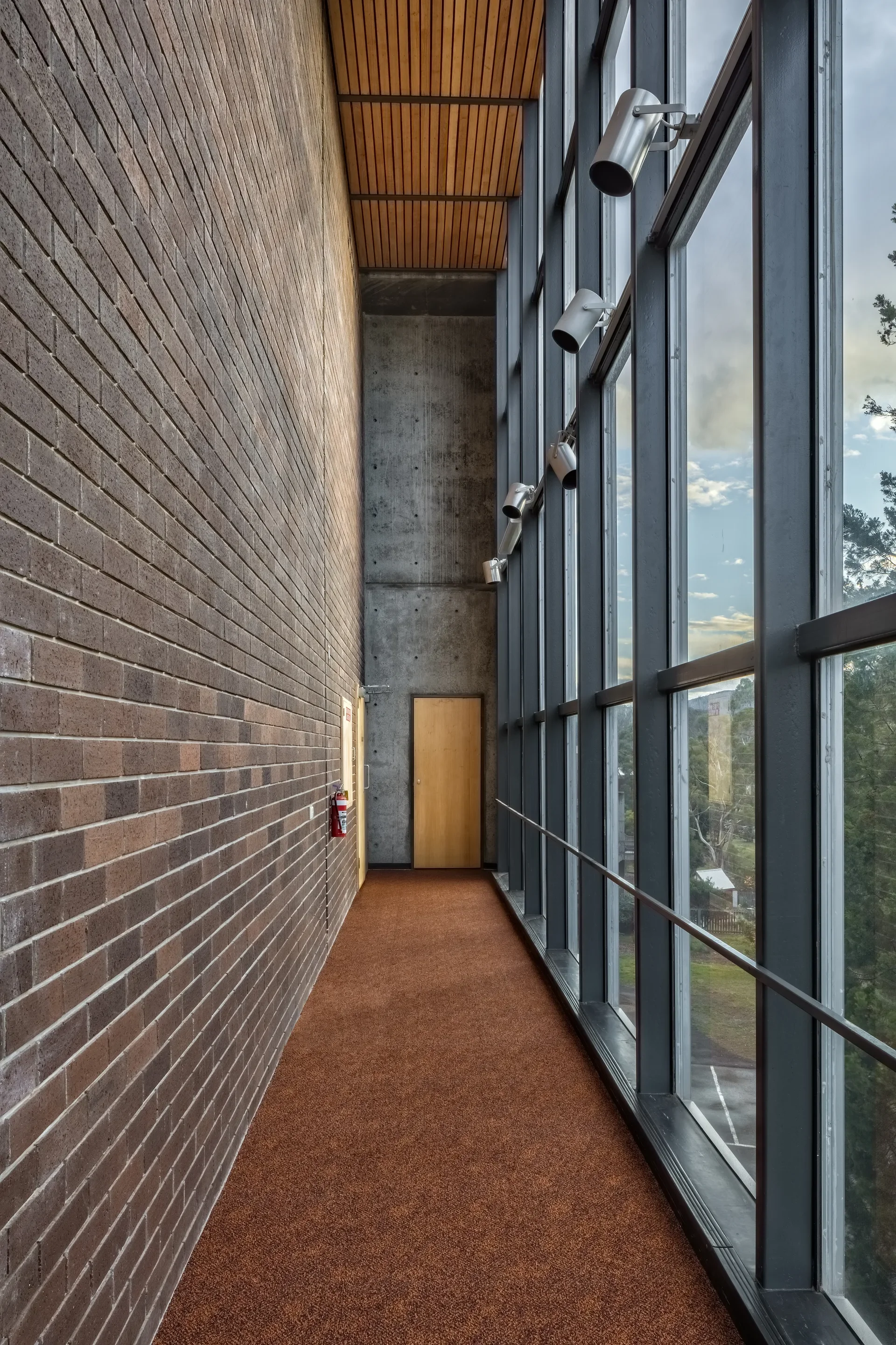 A narrow corridor features a brown carpeted floor, a dark brick wall, and a tall glass curtain wall lined with silver spotlights. At the far end, a wooden door is set into a concrete wall beneath a timber-slat ceiling.