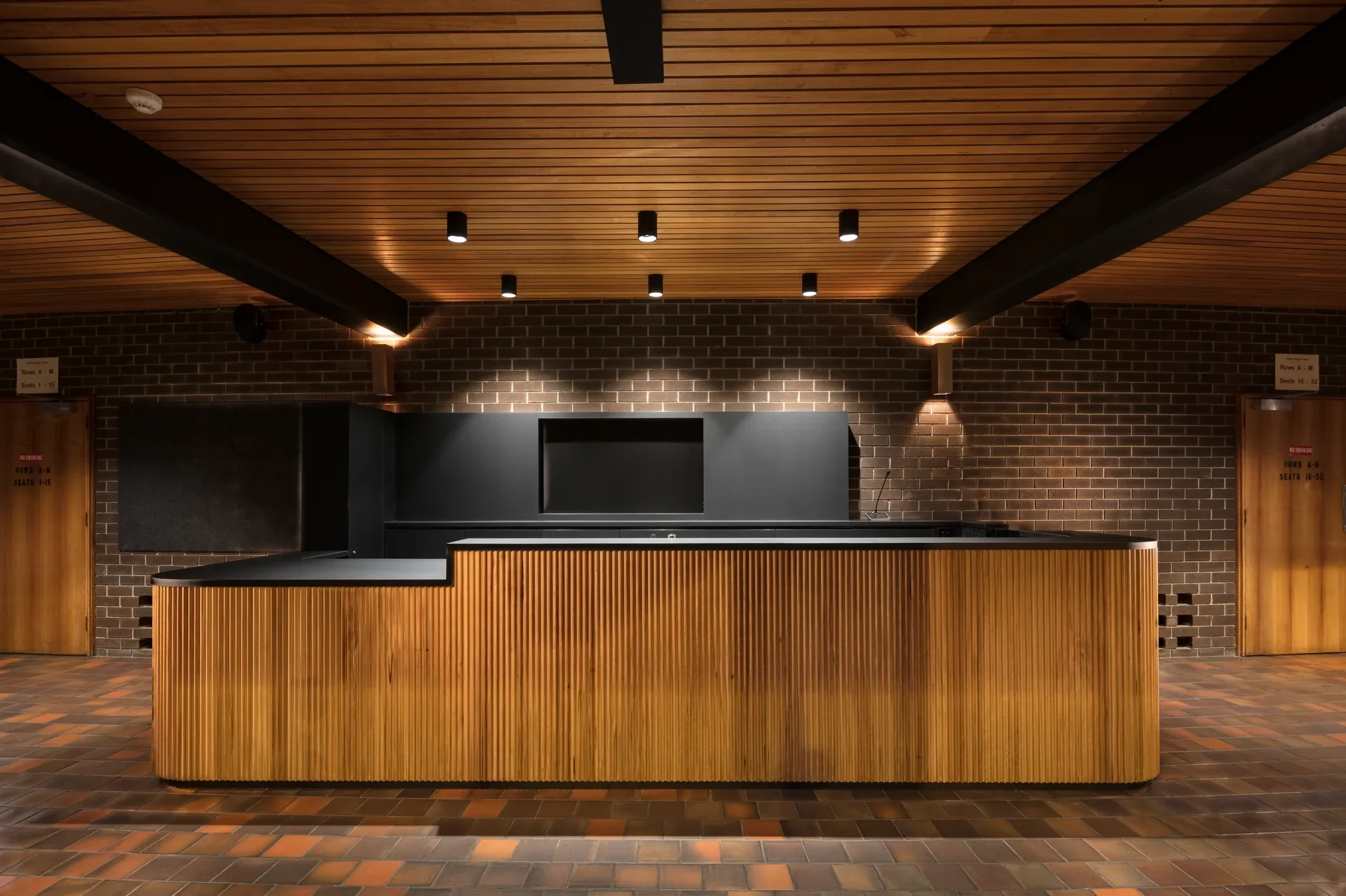 A timber-slat reception desk with a black top sits on terracotta tiles against a dark brick wall. The space features a timber-slat ceiling with recessed black spotlights and two wooden doors.