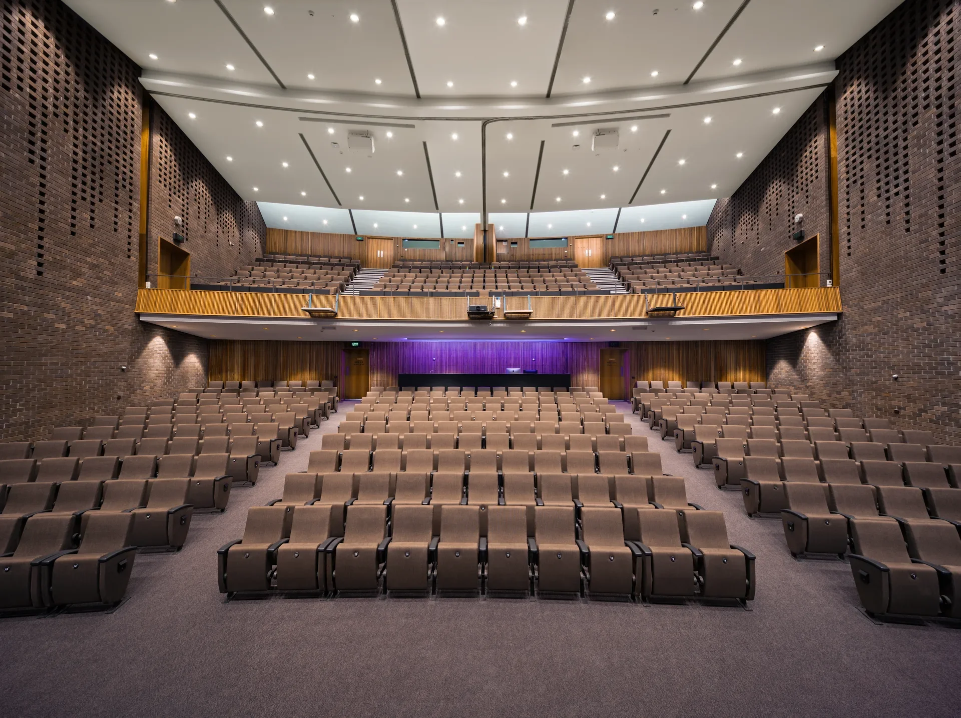 This large, tiered auditorium features brown upholstered seating, in the back some seats are illuminated by a purple glow. The space is defined by textured brick side walls, vertical timber-slat paneling, and a prominent wooden balcony level.