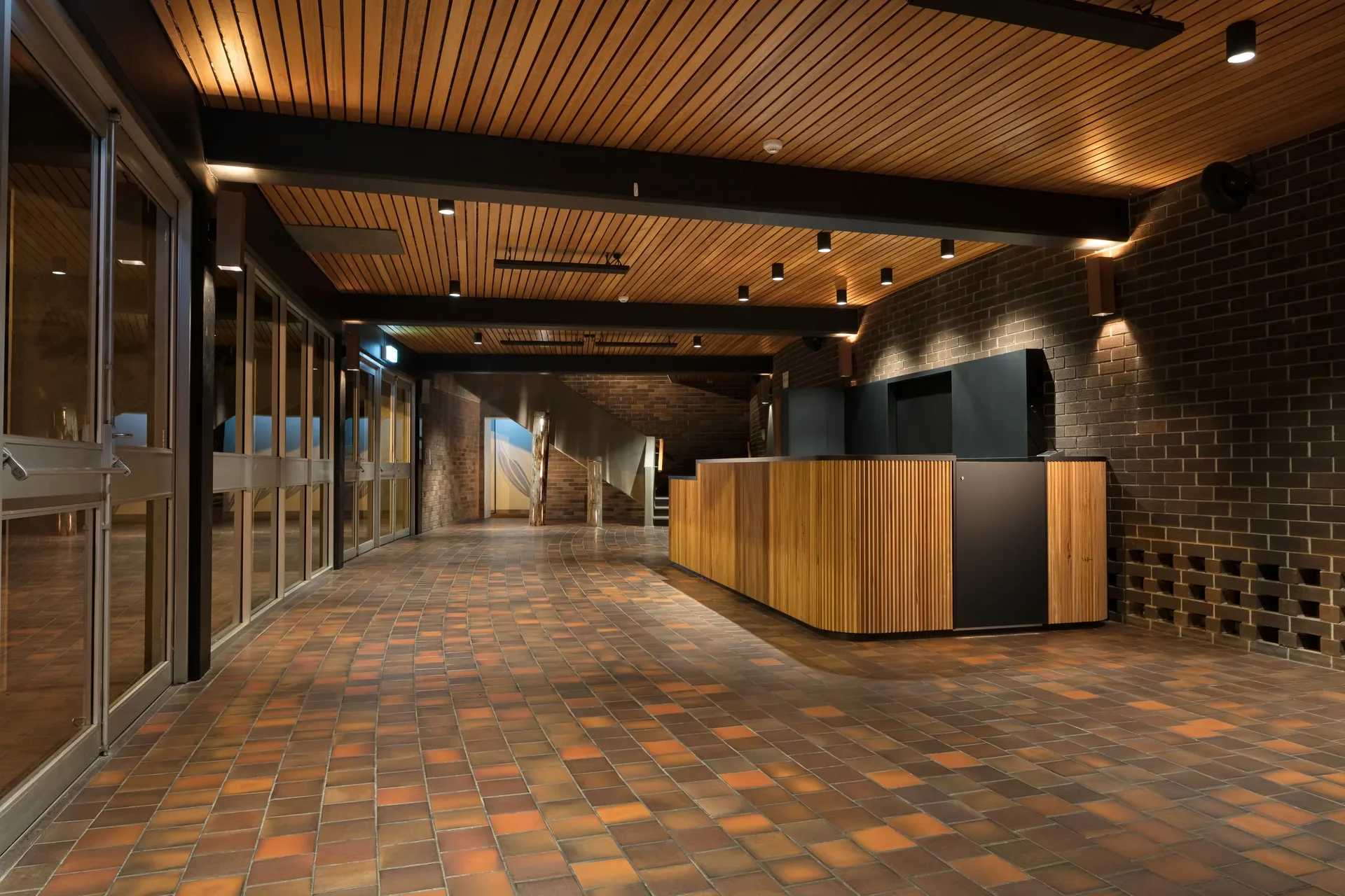 A building foyer features terracotta floor tiles, a timber-slat ceiling, and a wood-paneled reception desk against a dark brick wall. A series of glass doors on the left leads to an outdoor area.