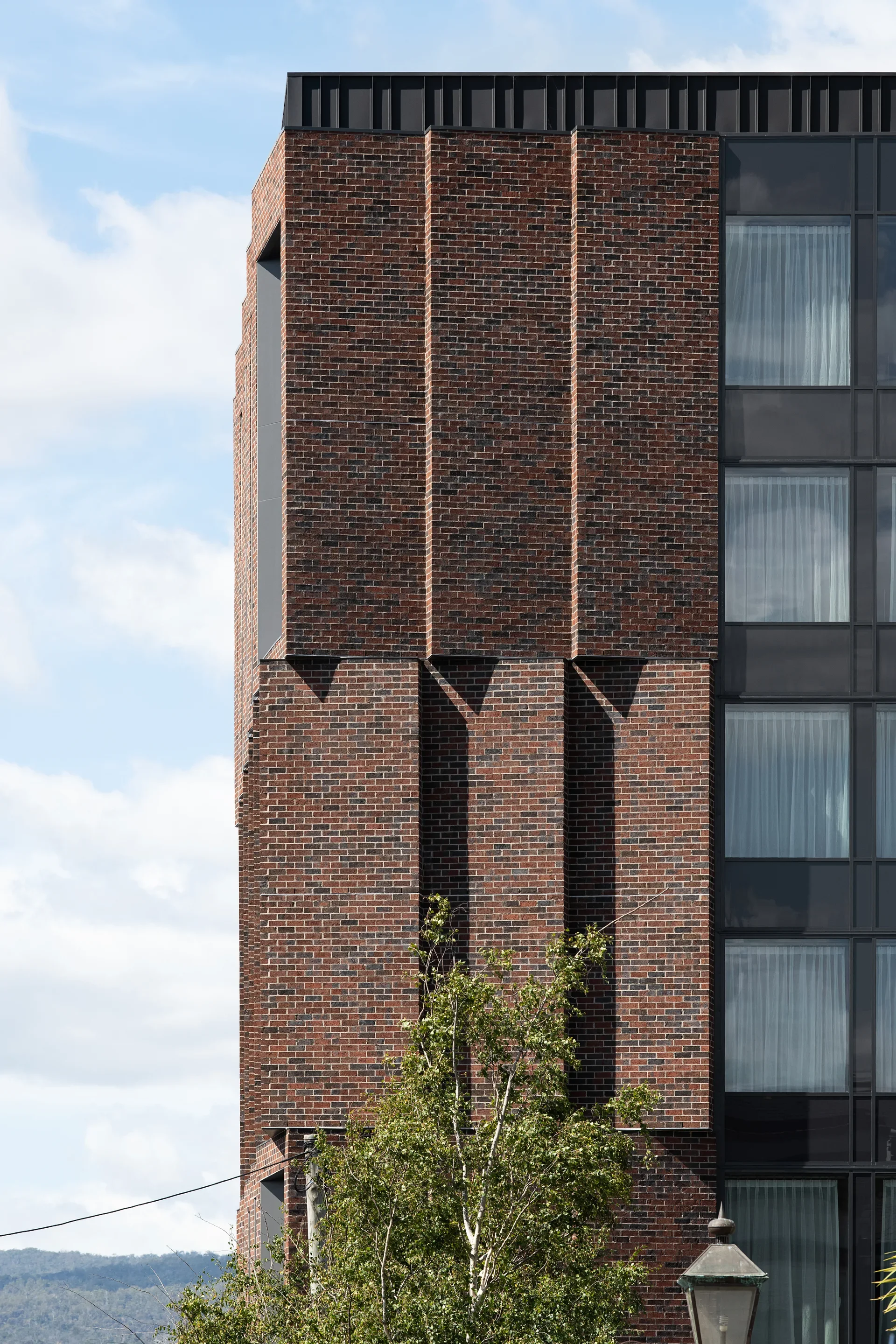 A red brick facade features vertical, angled walls that project deep shadows below next to a modern glass wall with dark metal framing. A leafy tree and a vintage lamp post stand in the foreground against a cloudy sky.