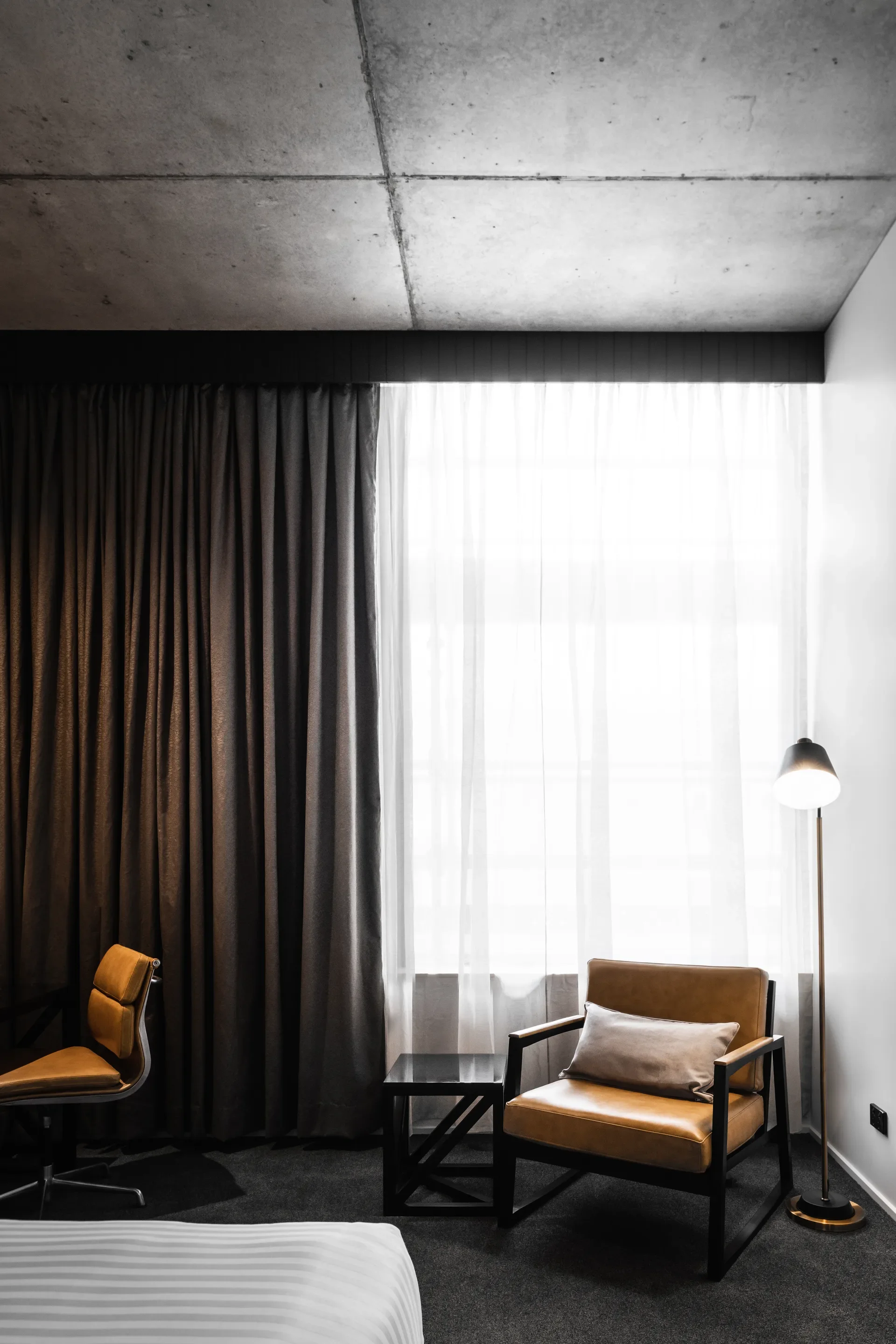 A modern bedroom features a concrete ceiling, dark curtains, and a tan leather armchair. The space is furnished with a small black side table and a slender floor lamp, all set against a large window with white sheer drapes.