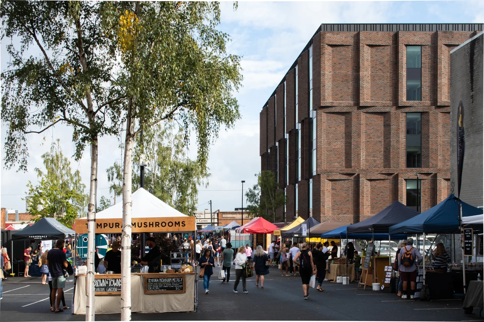 A bustling outdoor market features various food stalls, set against the backdrop of a modern red brick building. The brick facade has a distinctive sawtooth design, while birch trees and a crowd of people fill the foreground.