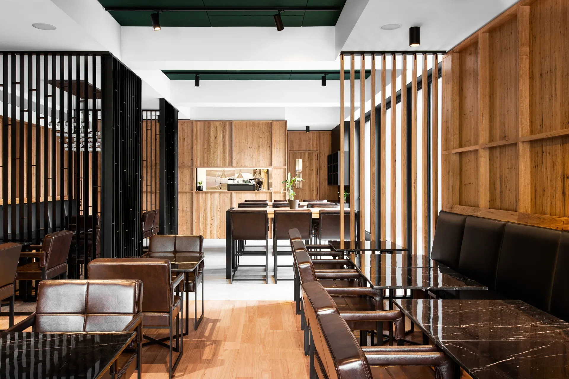 A modern dining area features timber-panelled walls, black vertical slats, and dark marble tables with brown leather seating. The space is lit by black spotlights against a green and white ceiling.