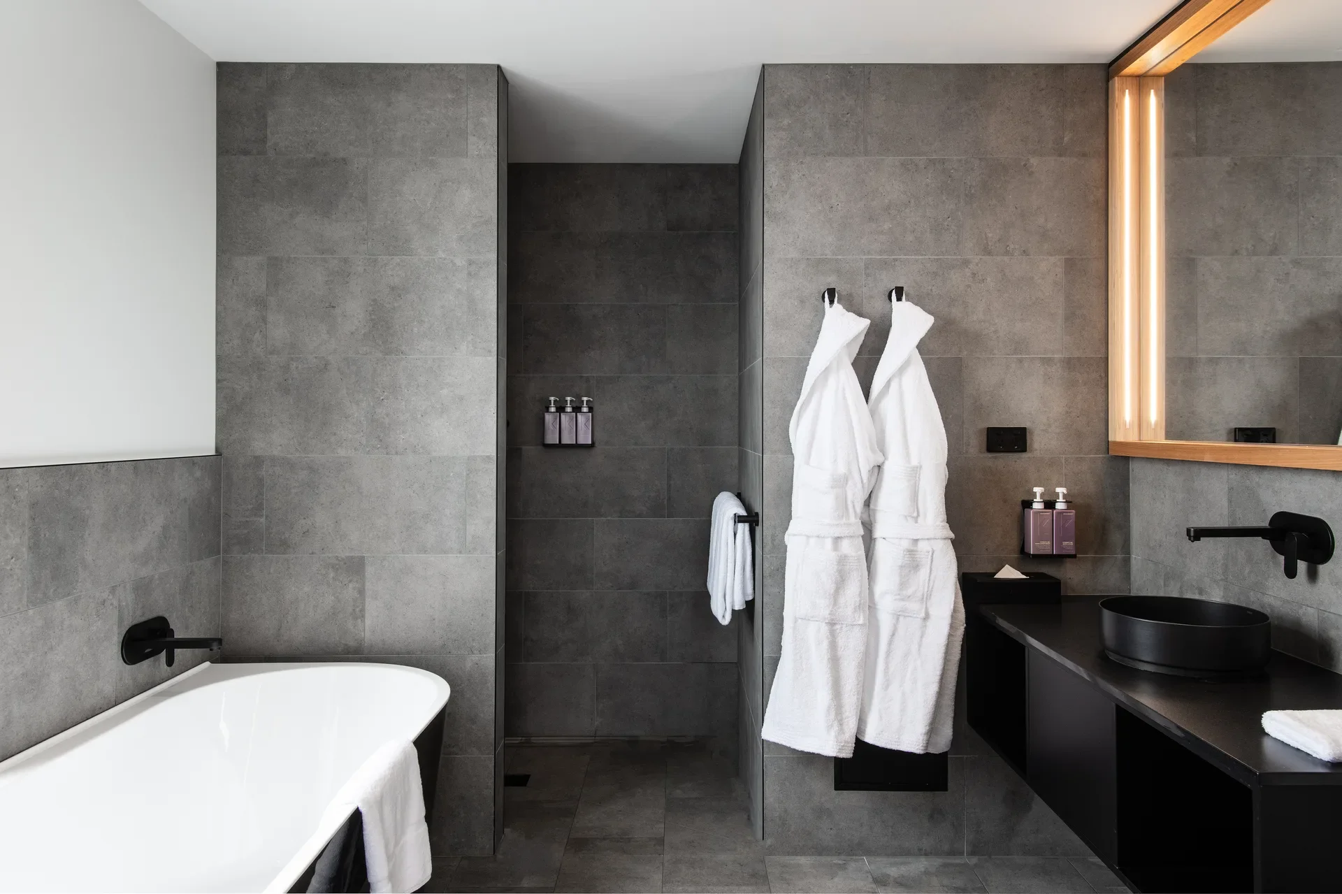 This modern bathroom features grey stone-textured walls, a white bath tub, and two white bathrobes hanging by a walk-in shower. A black vanity with a matching sink is illuminated by a vertical LED-strip mirror.