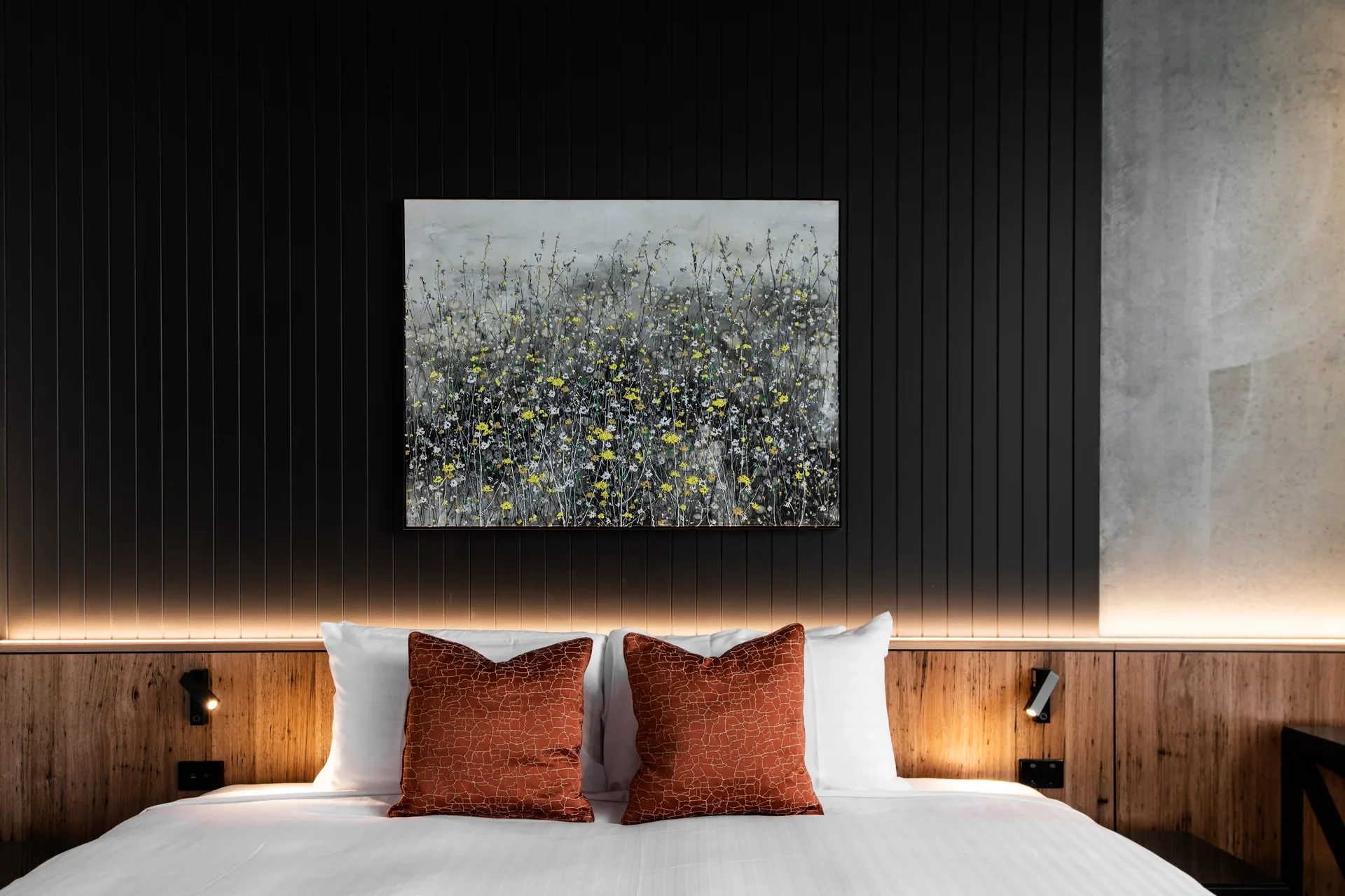 A floral painting hangs on a dark slat wall above a bed with terracotta pillows. The timber headboard features integrated reading lights and a warm under-shelf glow.
