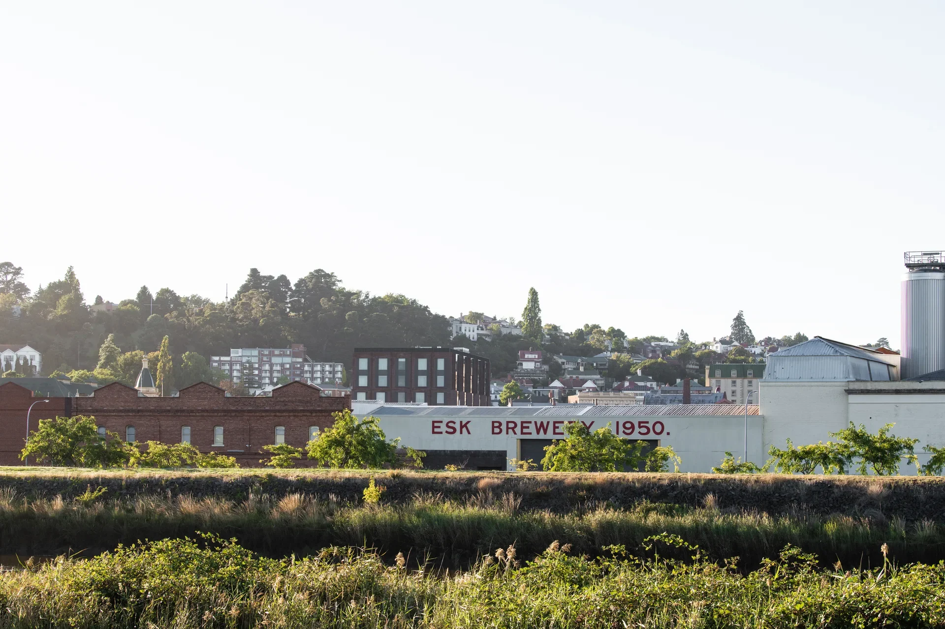 The red brick building stands amongst a city scape. In the foreground the "Esk Brewery" and its large white facade line the banks of the Tamar River.
