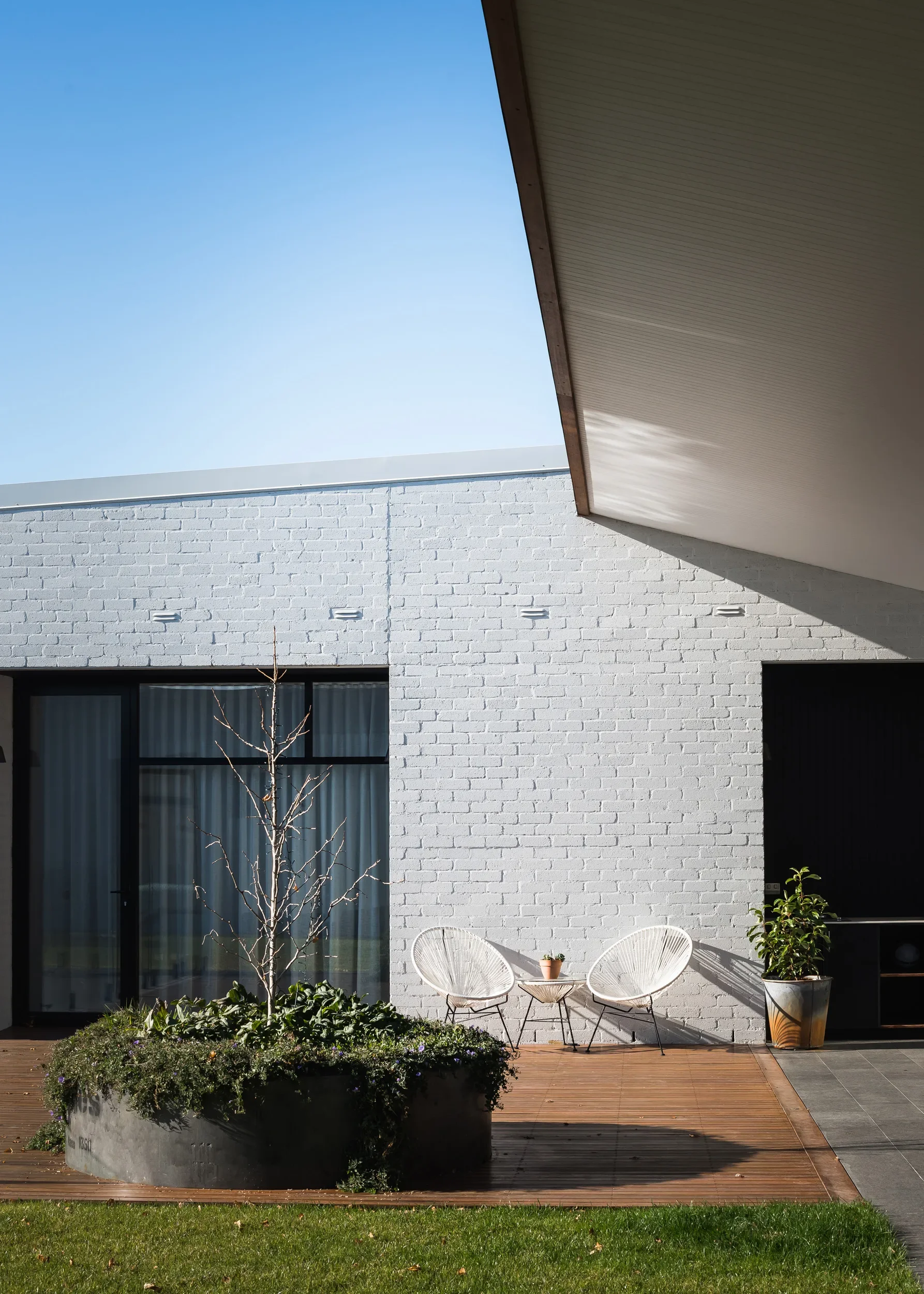 A sunlit patio features a white brick wall, a wooden deck, and two white chairs with a small side table. A large circular planter sits in the foreground beneath the shadow of an overhanging roof.