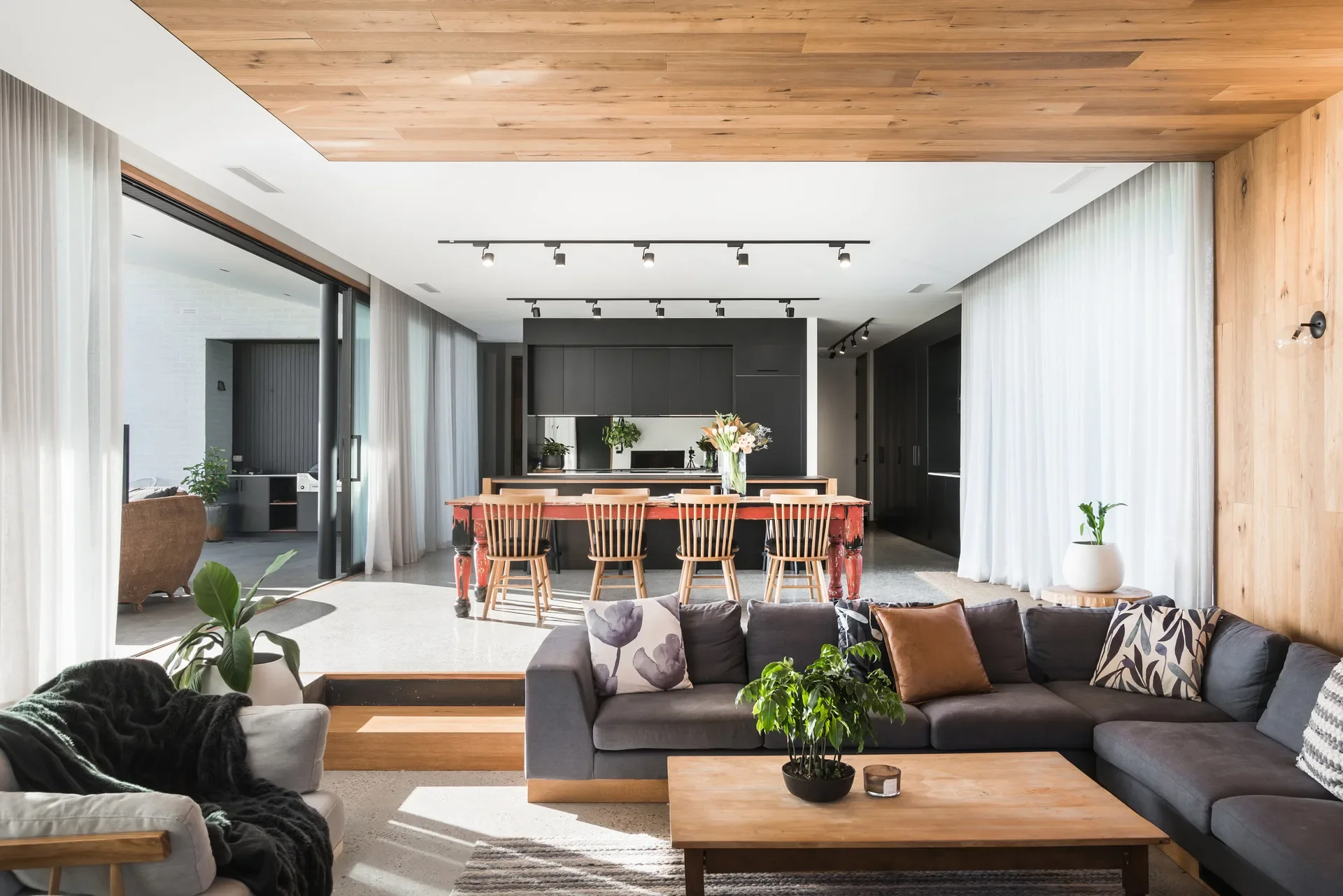 This open-plan space features a wood-panelled ceiling, a grey sofa, and a red rustic dining table. A sleek black kitchen stands in the background, while glass doors open to a sunlit patio.