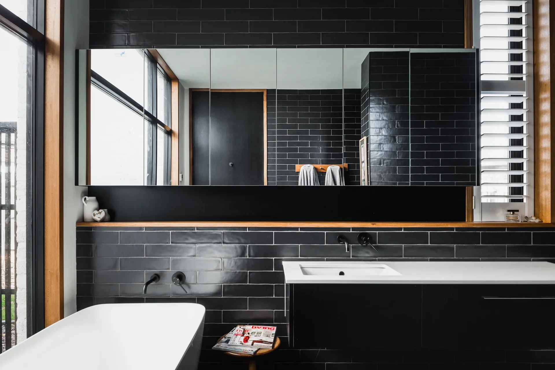 This modern bathroom features black brick-style tiles, a white vanity, and a large mirrored cabinet. A white freestanding tub is visible in the foreground next to a small wooden stool.