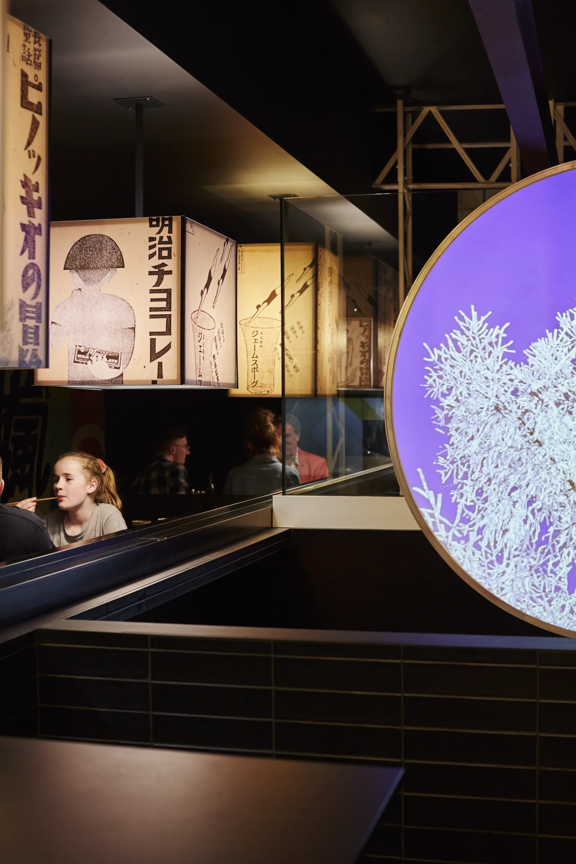 A dimly lit restaurant features Japanese-style light box lanterns and a large circular purple light with a tree-like pattern. People are seated at dark dining booths in the background.