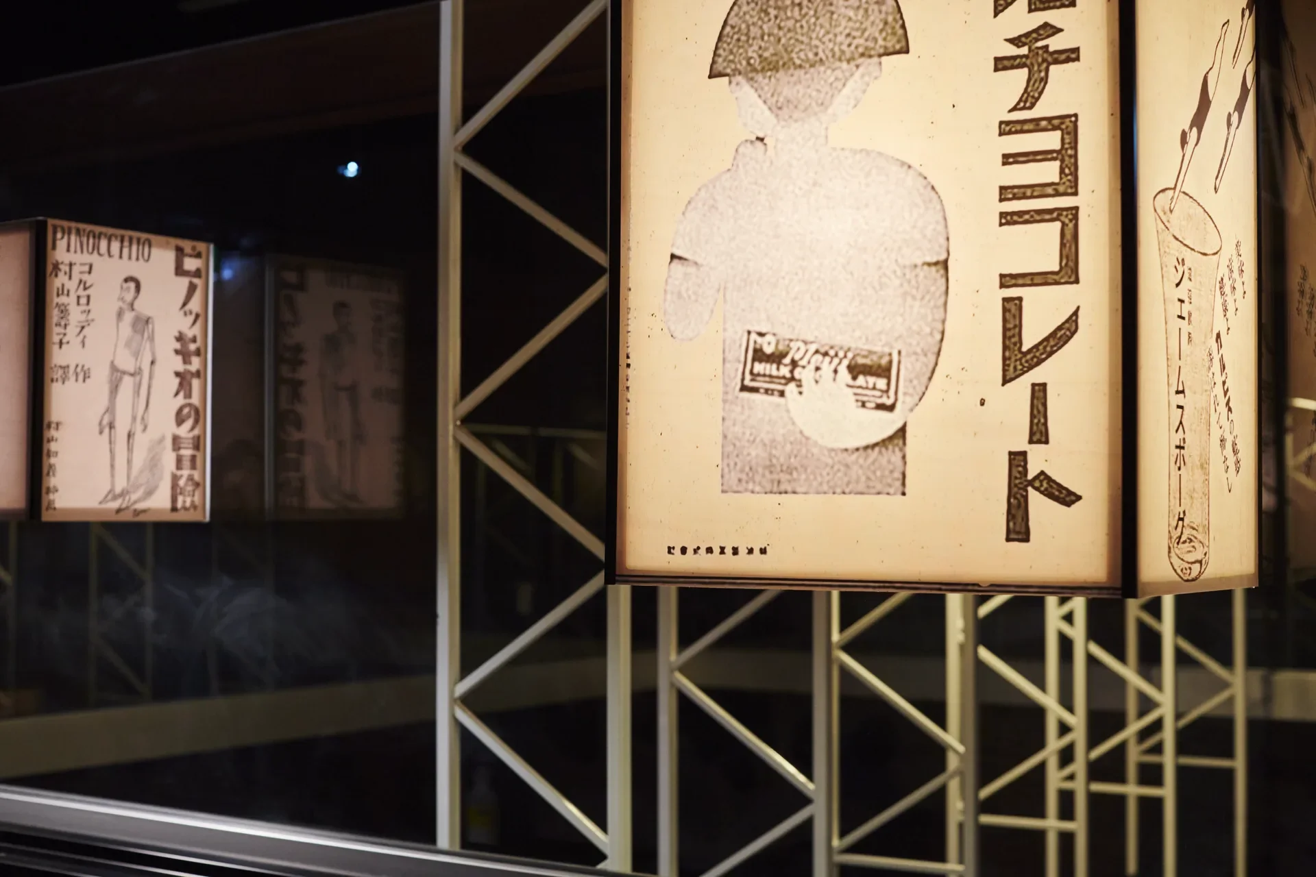 Japanese-style light box lanterns featuring black-and-white graphics and vertical text hang against a dark background. A white steel frame is visible behind the lanterns.