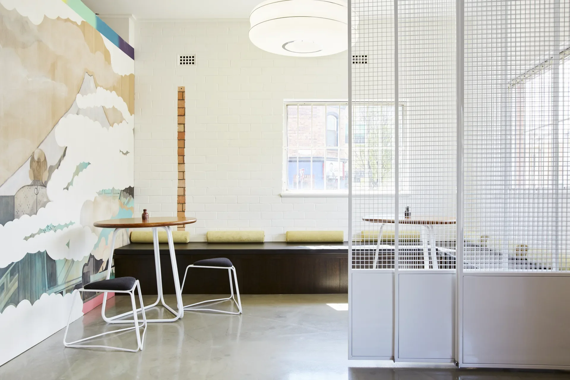 A cloud and mountain mural decorates a white brick wall next to a dark bench with yellow bolsters. The bright space is divided by a white mesh screen and furnished with round wooden tables.