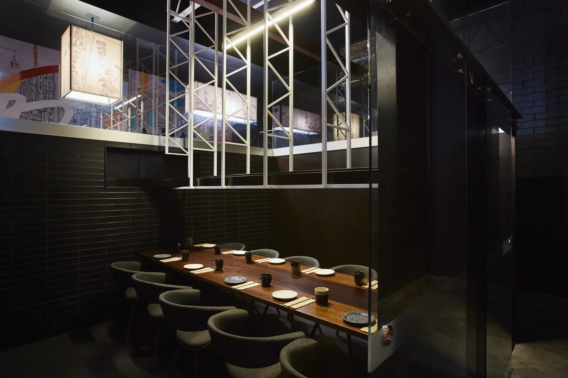 This private dining area features a long wood table set for eight with curved grey chairs. A white geometric framework hangs overhead, surrounded by dark tiled walls and a glass partition.