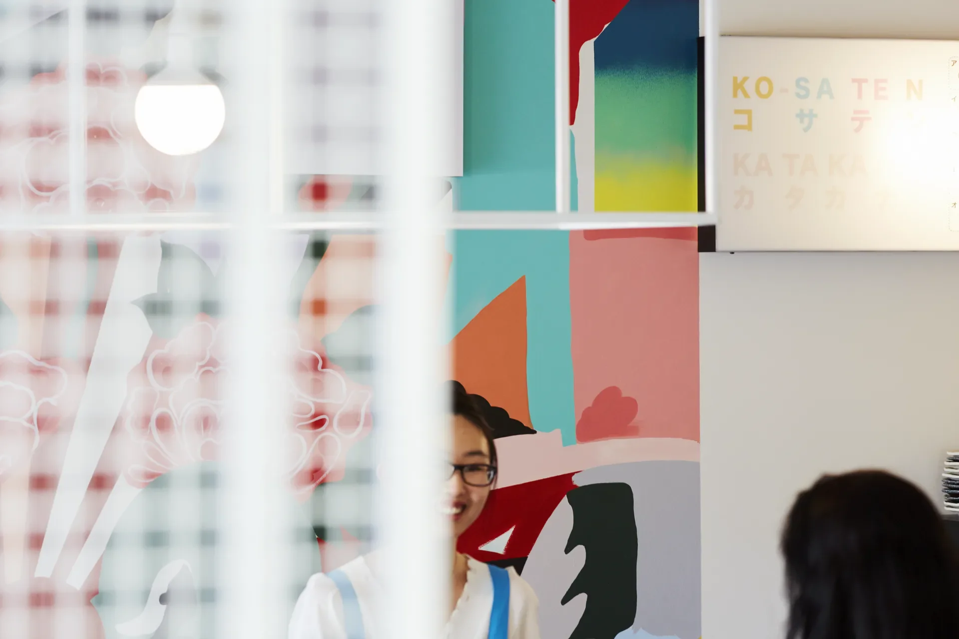 A vibrant multicolored mural is visible behind a white geometric framework and a frosted glass screen. A staff member stands near a sign reading "KO-SA-TE-N".