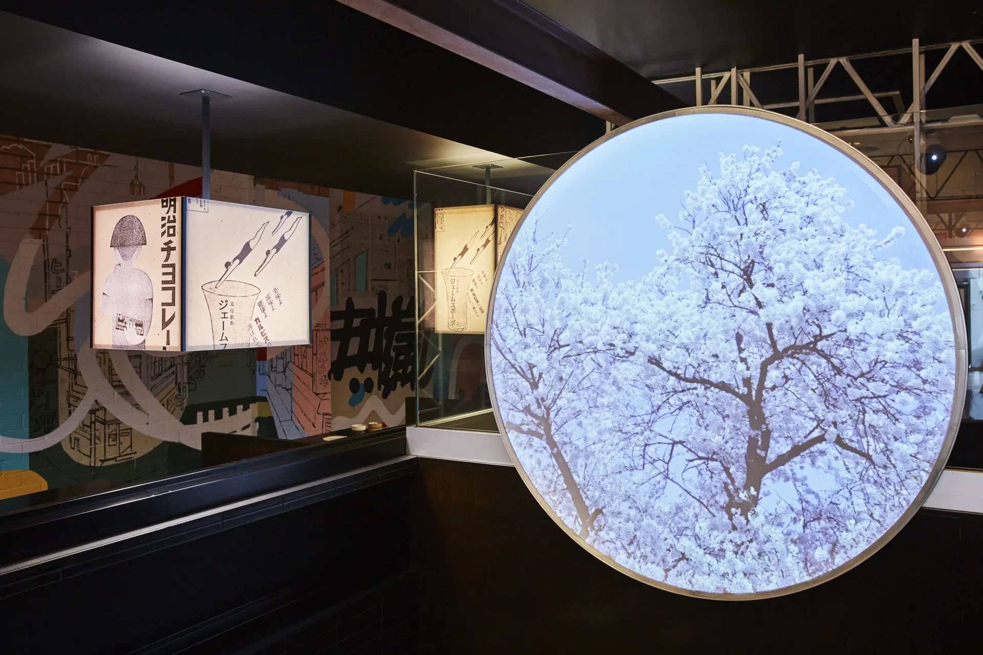 A large, circular light box featuring a white cherry blossom tree glows next to a Japanese-style lantern with black and white illustrations. Colourful murals and dark partitions are visible in the background.
