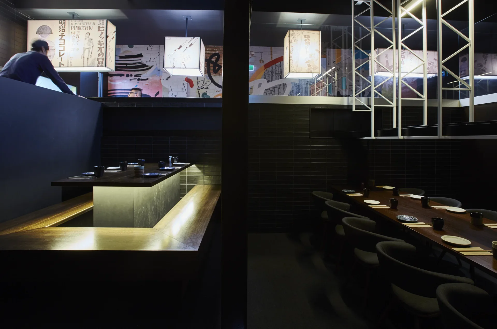 This restaurant features sunken-style seating with glowing under-lighting on the left and a traditional long wooden table on the right. Japanese-style light box lanterns and a white geometric framework hang above the dining areas.