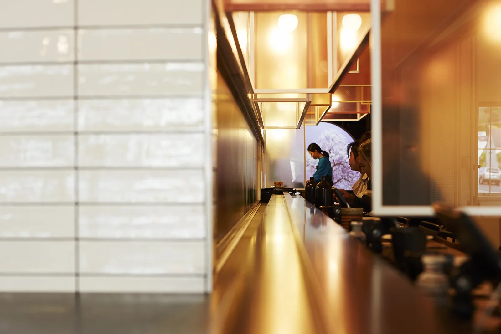 A long, polished counter reflects warm light in a narrow restaurant where customers are seated. In the background a large purple circular glowing light.