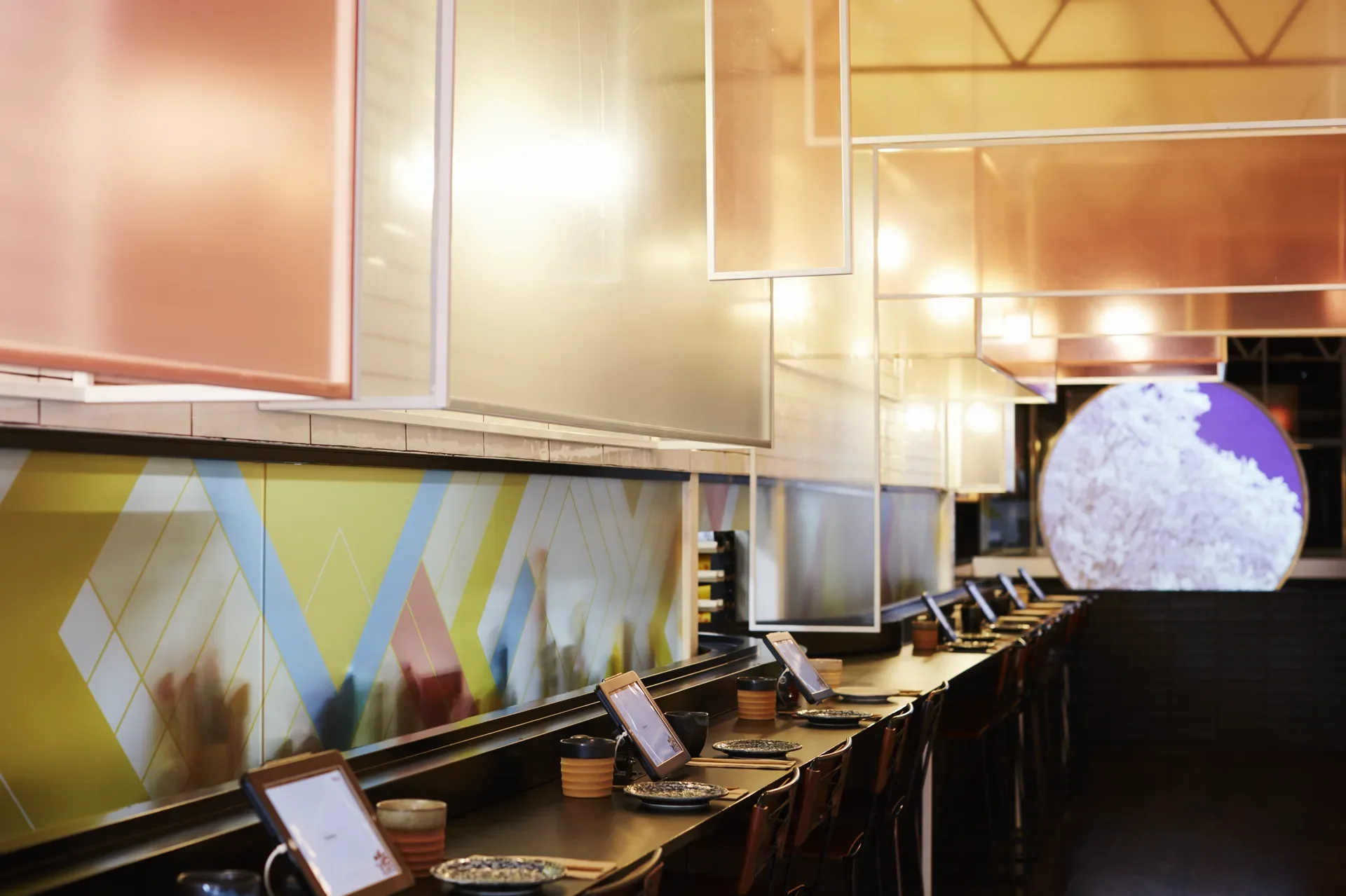 This modern dining area features a long dark counter with tablet ordering screens and minimalist place settings. Translucent orange and white panels hang overhead, while a large circular light with a tree pattern glows in the background.