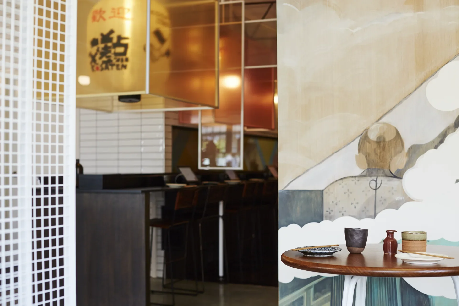 This dining space is divided by a white mesh screen and features a round wooden table set with minimalist crockery. Translucent orange panels and a large yellow lantern with Japanese text hang above a dark counter in the background.