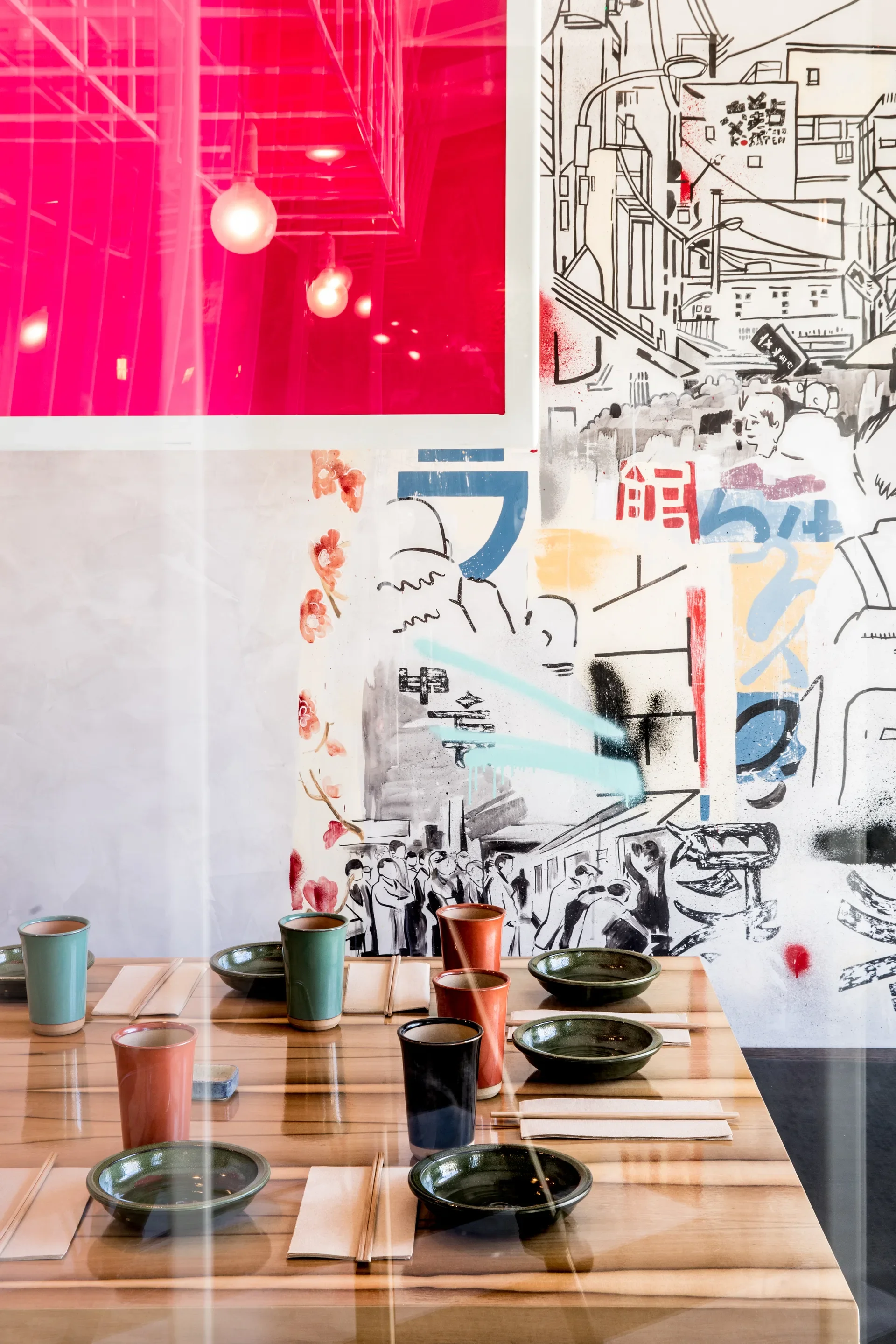 A wooden table is set with dark green bowls and colorful ceramic cups. The background features a graffiti-style mural and a large vibrant pink panel reflecting overhead lights.