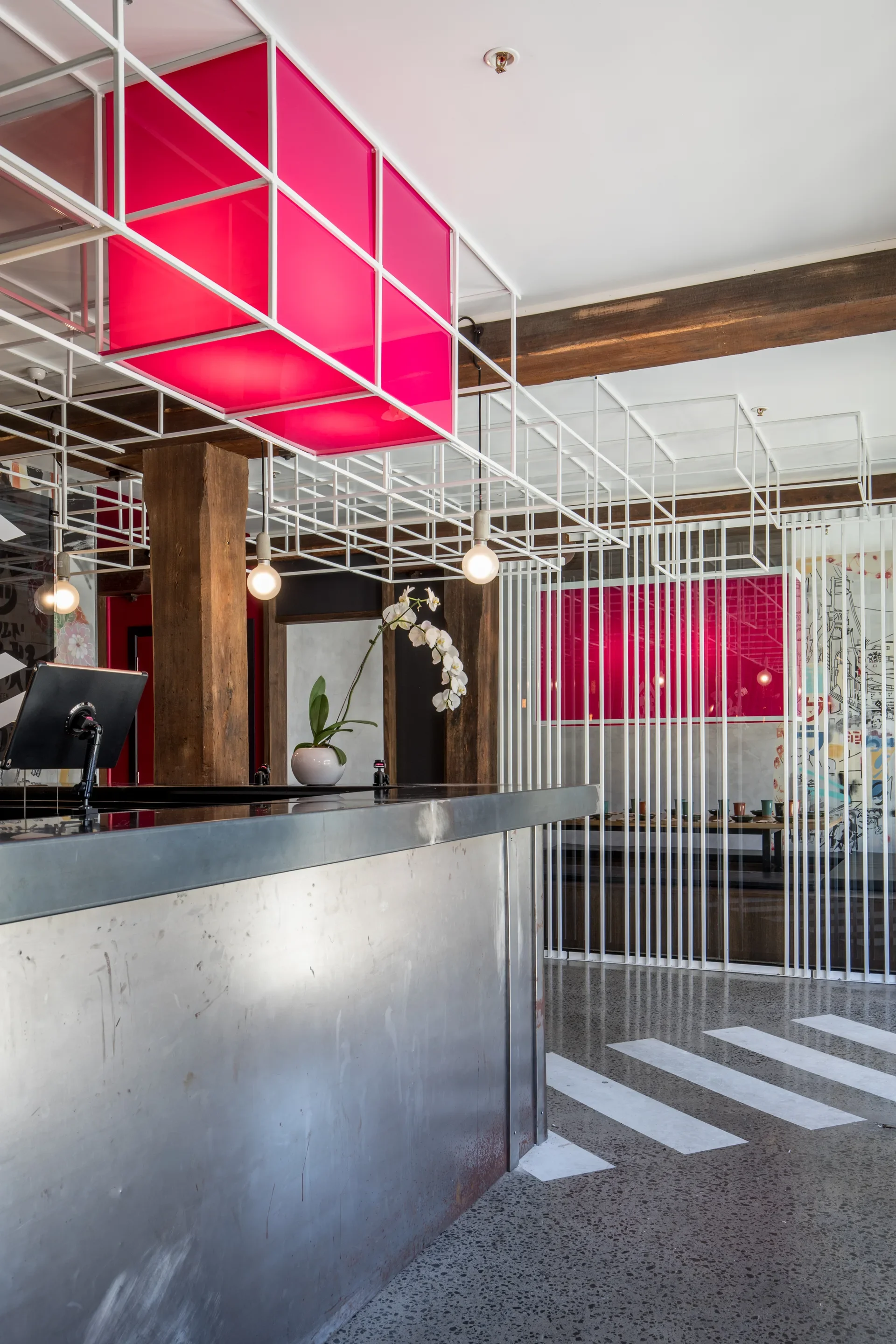 This industrial space features a metallic service counter beneath a white ceiling grid holding a vibrant pink light box. White diagonal stripes mark the polished floor, while vertical slats separate the counter from the dining area.