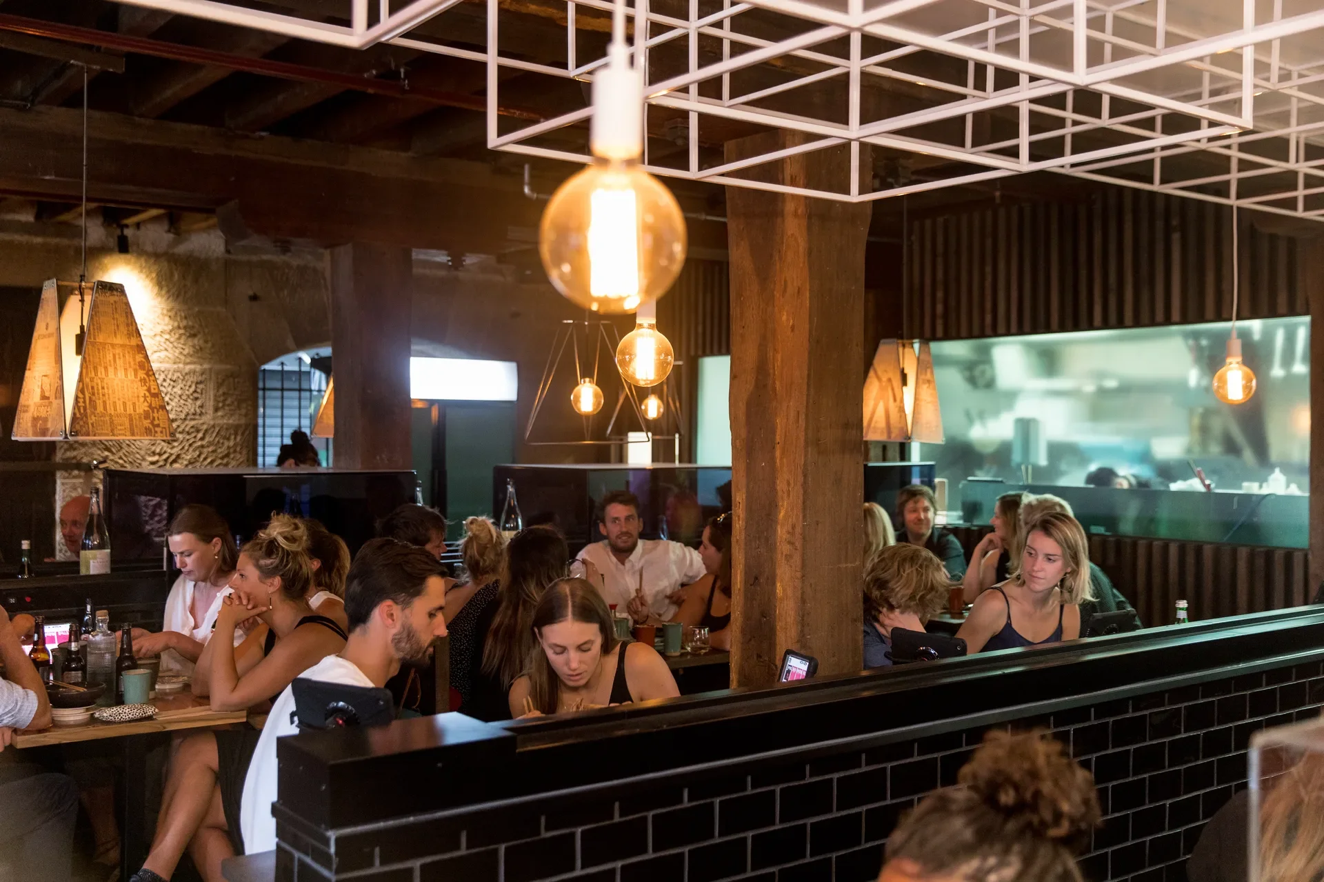 This crowded restaurant features customers dining beneath pendant light bulbs and a white geometric ceiling grid. Thick wooden pillars and dark tiled walls divide the space, with the kitchen visible in the background.