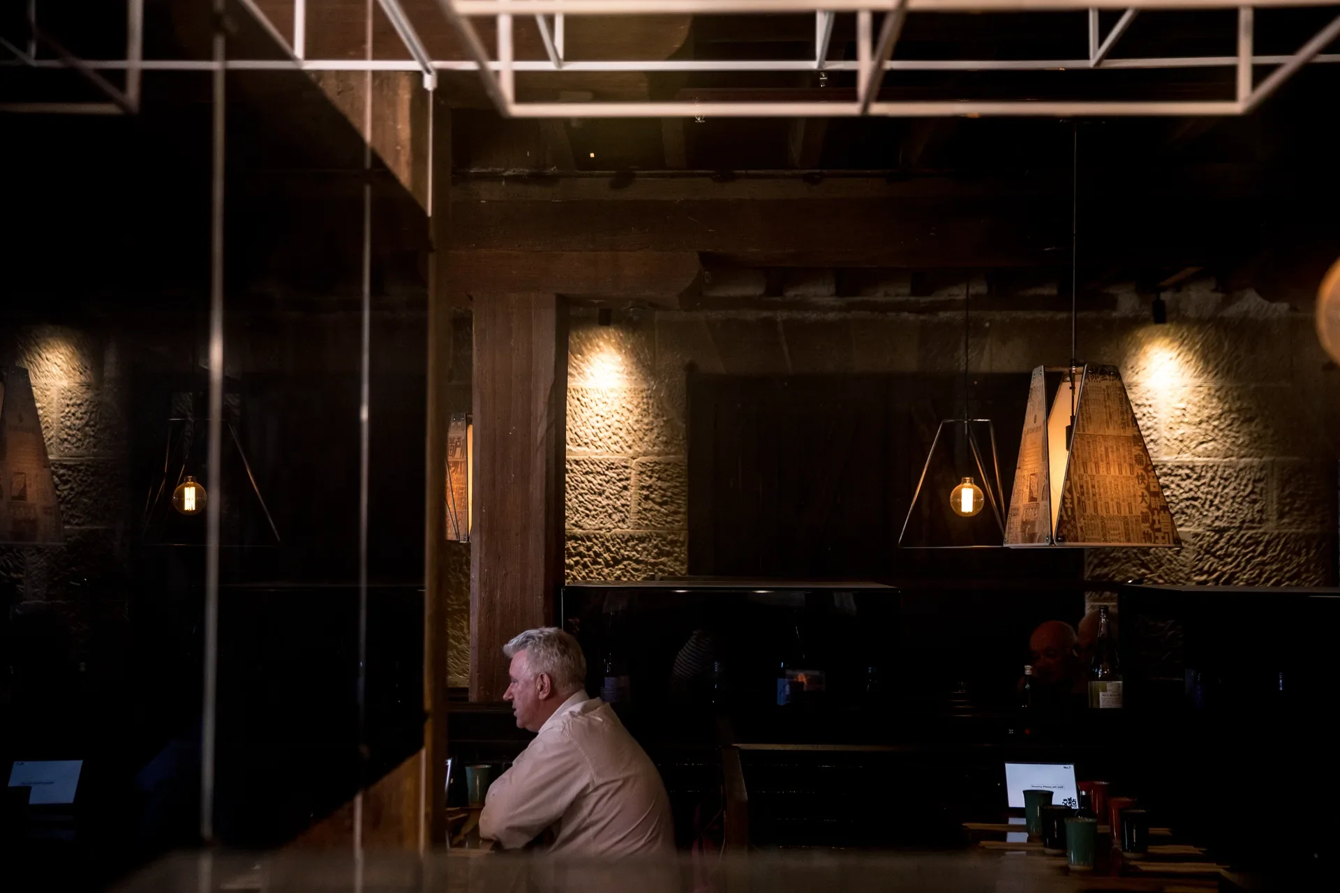 This dimly lit space features exposed stone walls, thick wooden pillars, and customers dining at tables. Geometric pendant lights and lanterns with newspaper-style graphics provide the primary illumination.