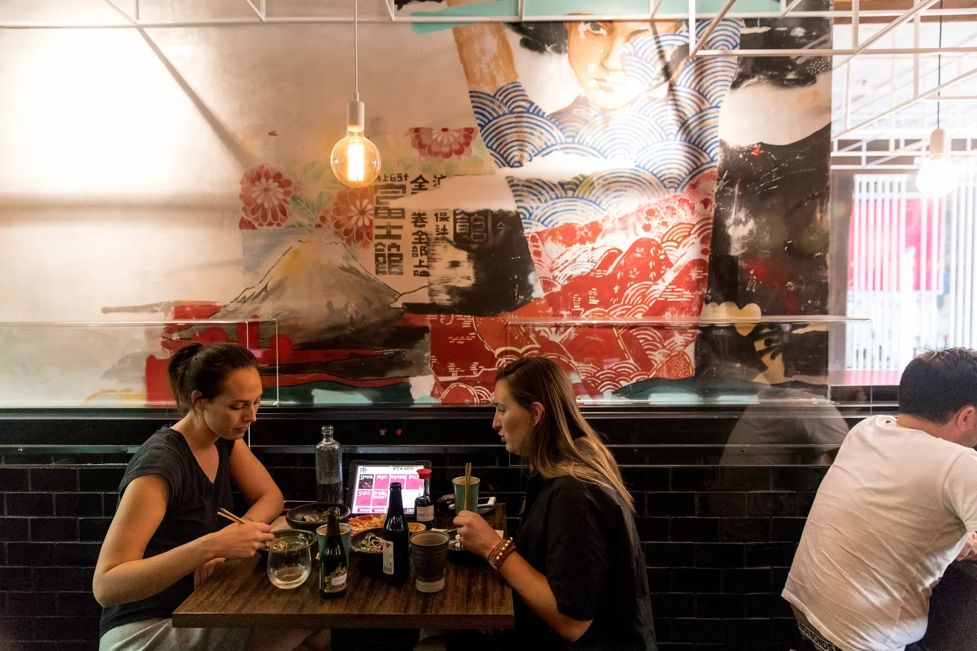 This dining area features a vibrant mural of a stylised figure and mountain behind customers at a dark table. A pendant bulb hangs from a white ceiling grid, and a digital tablet is used for ordering.