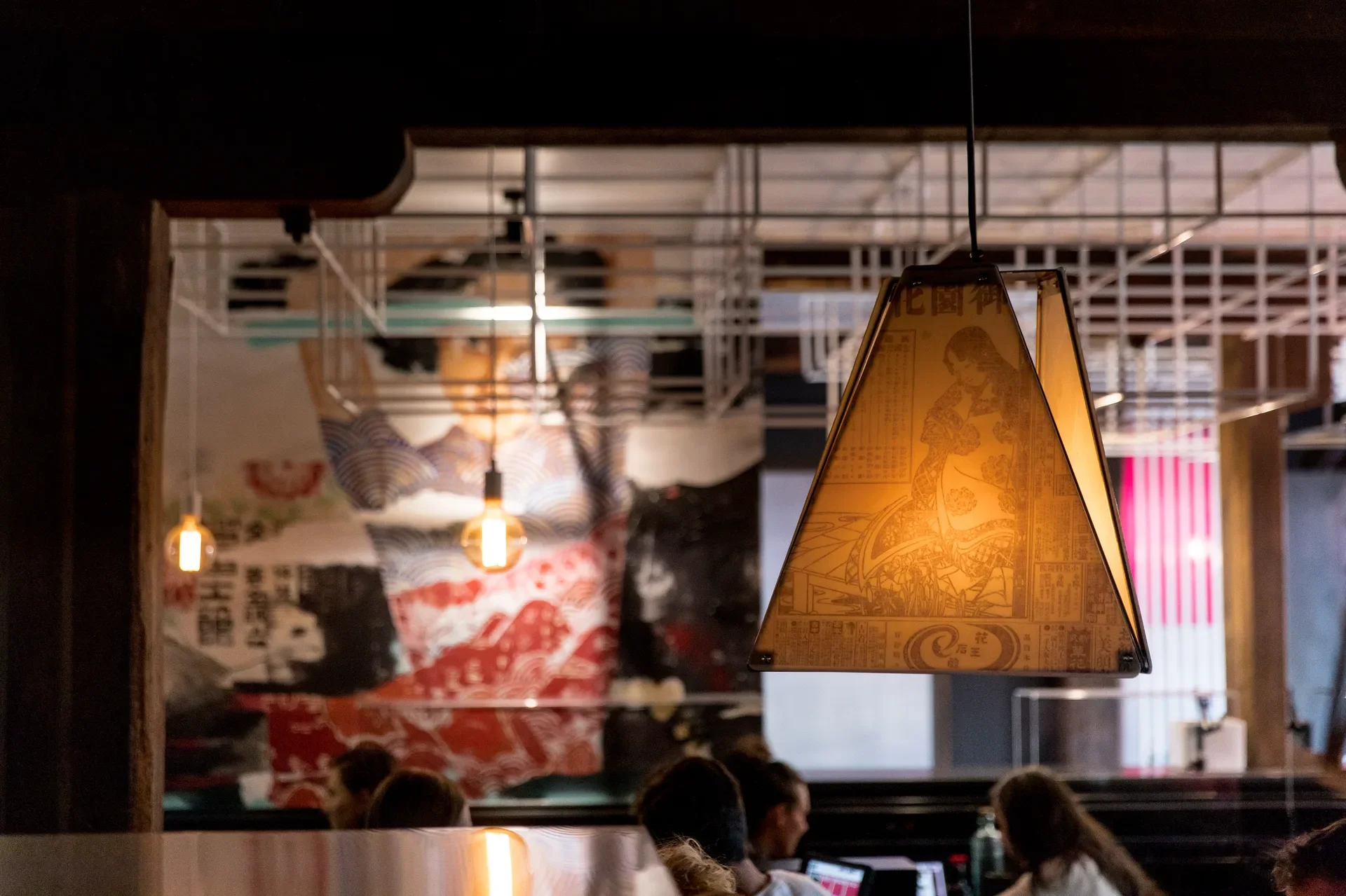 A trapezoidal lantern with Japanese graphics hangs in the foreground. A vibrant mural and white geometric ceiling grid are visible in the background.