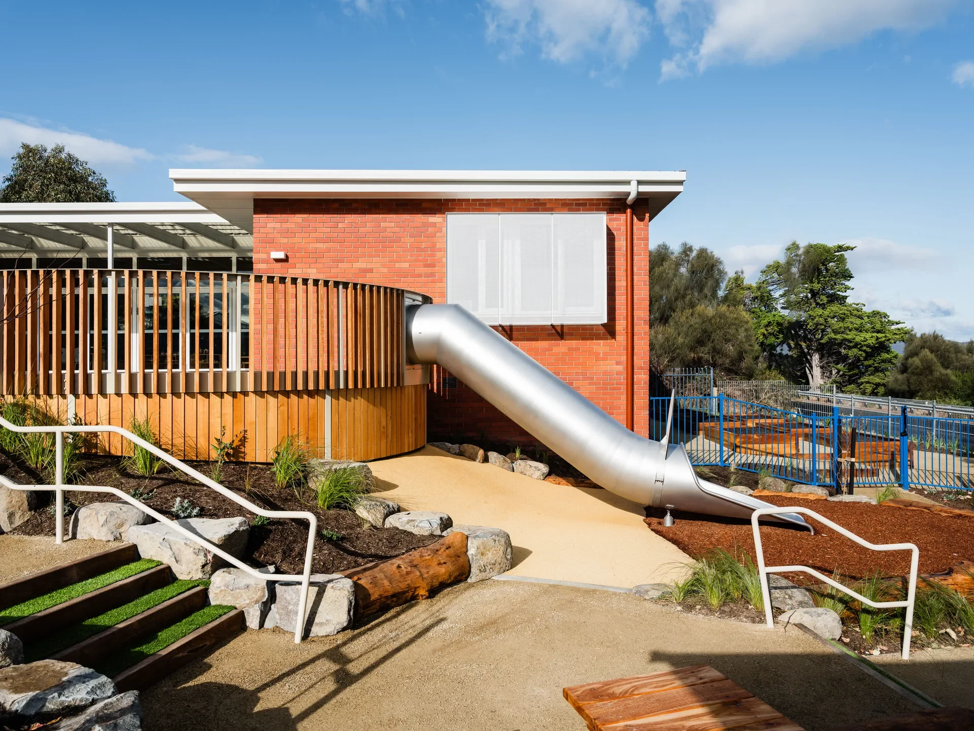 This outdoor area features a metal slide extending from a red brick building and a curved wooden deck. The landscape includes stone-lined beds and a staircase with green turf treads.