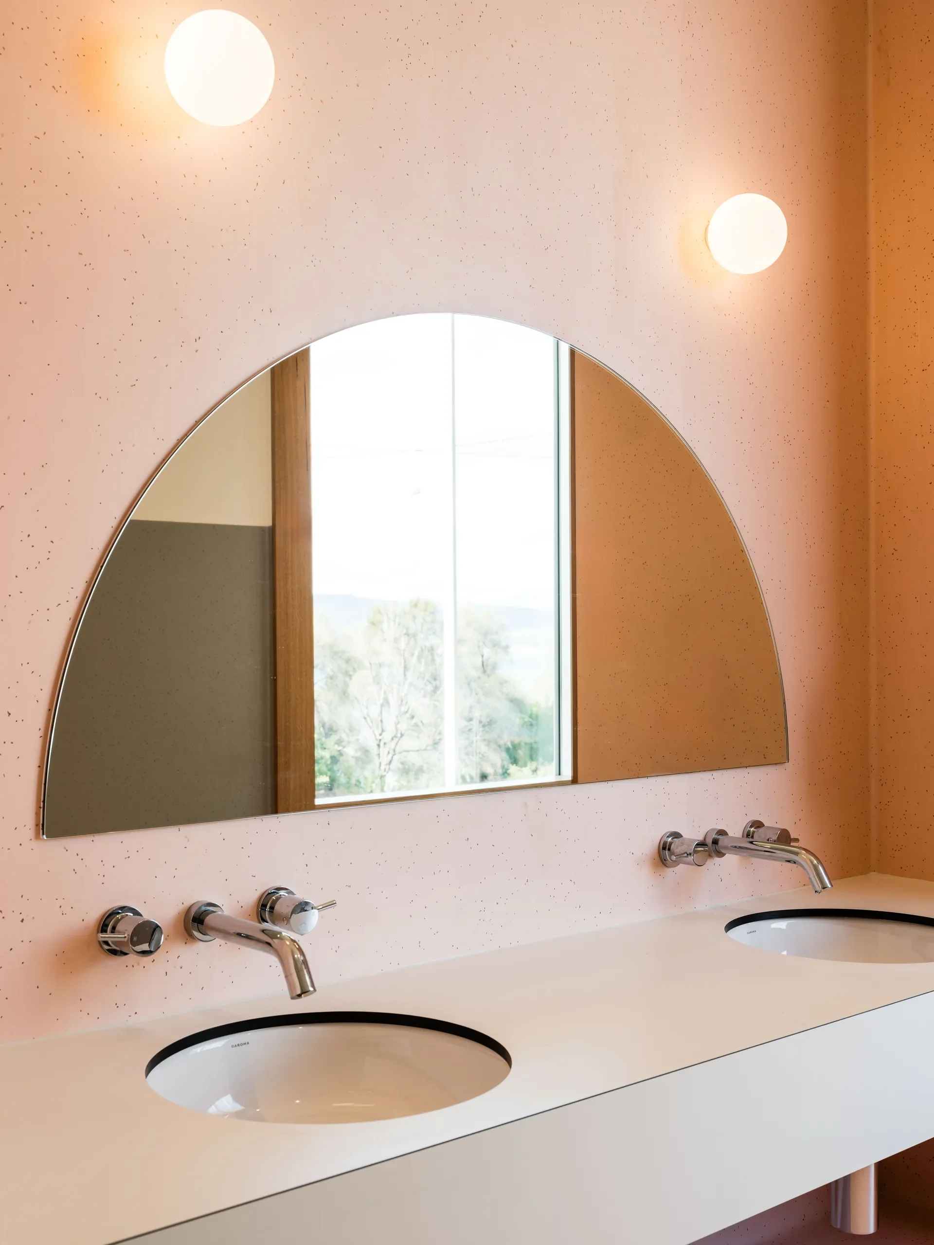 This minimalist bathroom features a semi-circular mirror on a speckled pink wall between two globe lights. A white vanity holds two sinks with modern silver faucets.