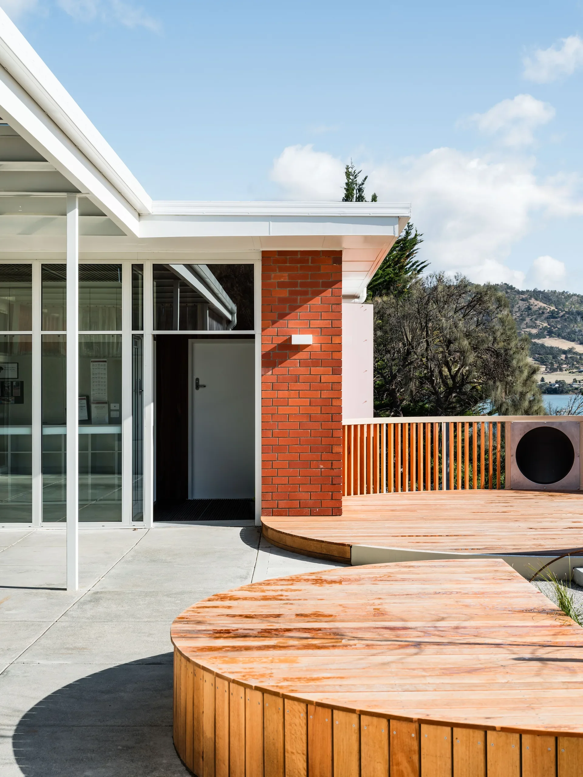 This building features a red brick wall, floor-to-ceiling glass windows, and a curved wooden deck. The background offers a scenic view of water and distant hills under a bright sky.