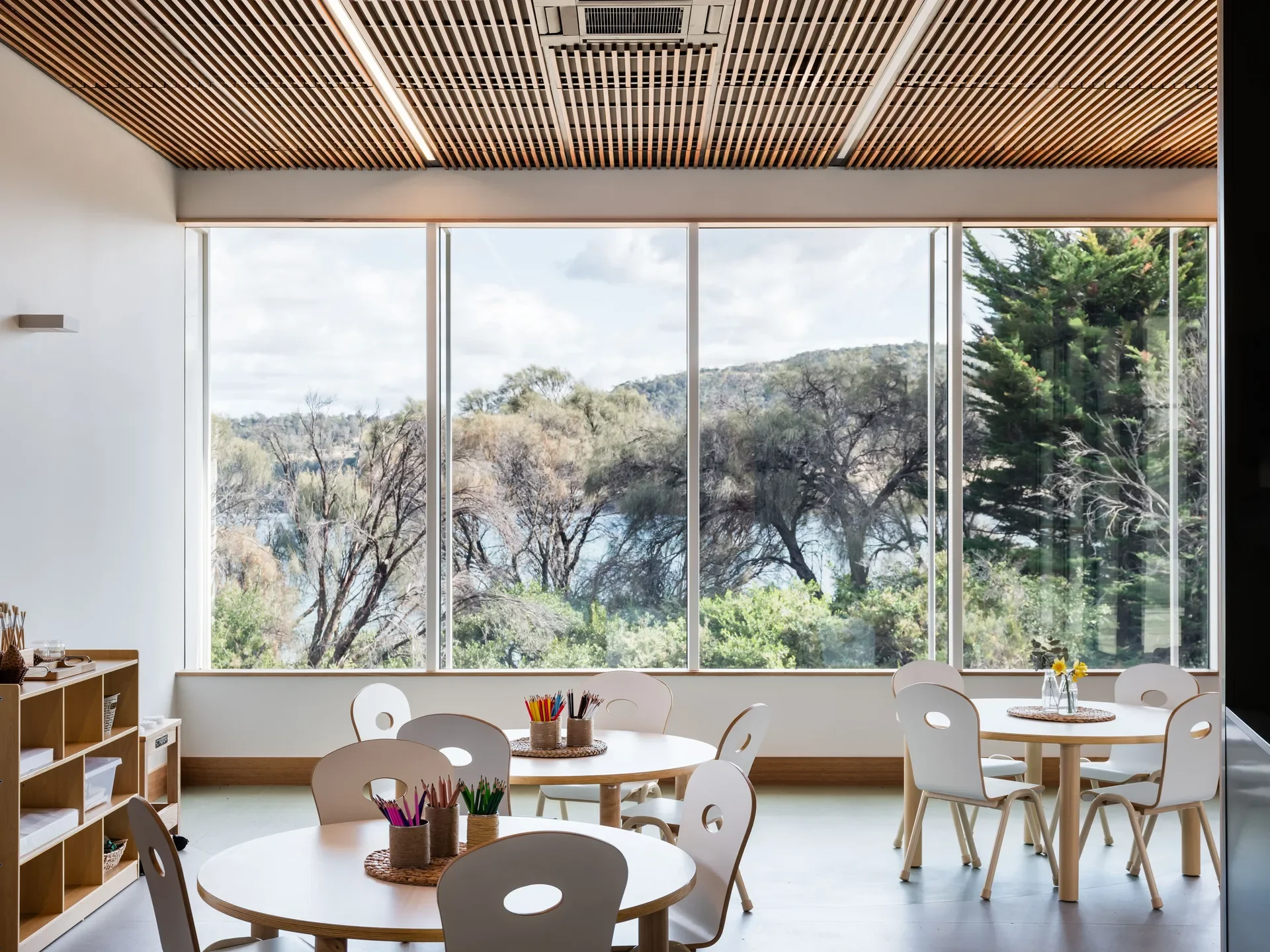 This bright classroom features slatted wood ceilings and large windows overlooking a lush landscape of trees and water. Several round tables with white chairs are set with cups of coloured pencils, while a low wooden shelf stands to the left.