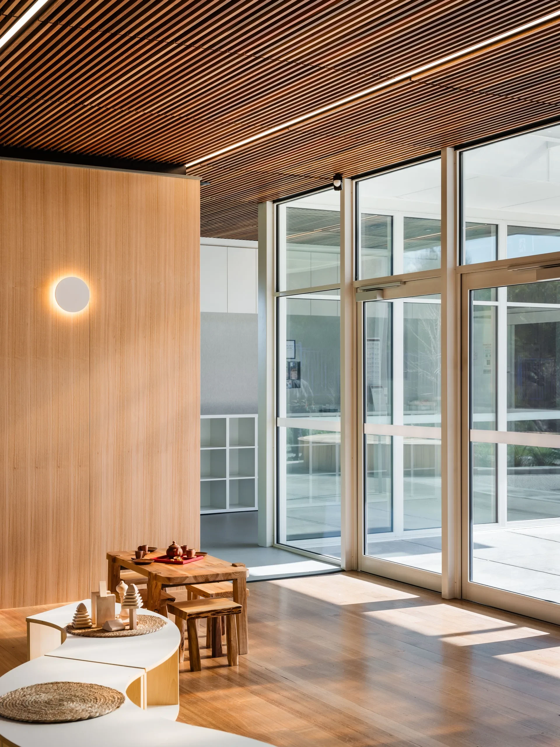 This sunlit room features slatted wood ceilings, a light-wood wall with a circular sconce, and large glass doors. Curved white shelving and a wooden tea table sit on the polished hardwood floor.