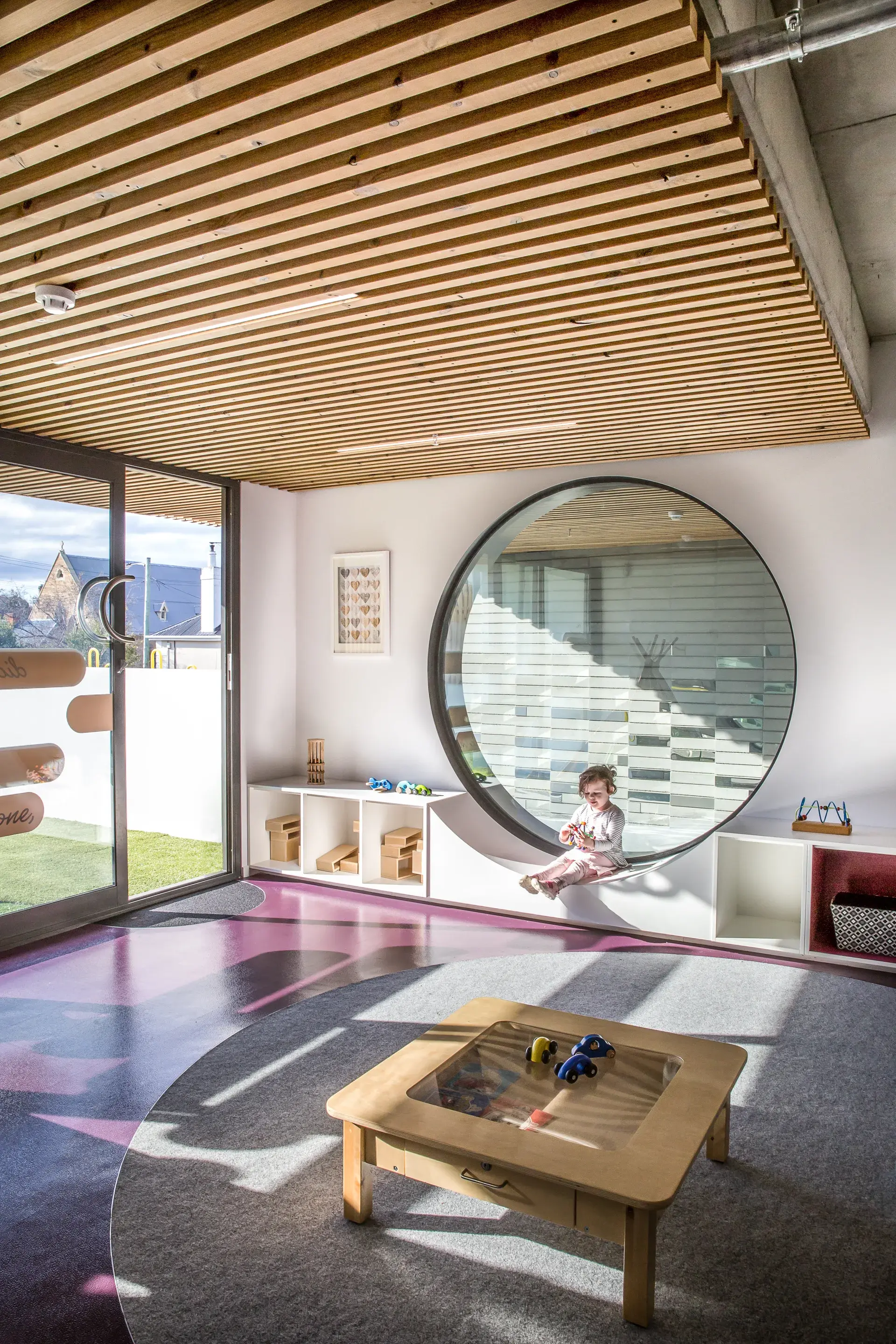 This bright room features slatted wood ceilings, a grey circular rug, and a large round window with a built-in seat where a child is sitting. Low white shelving and a small wooden activity table furnish the space, which opens to an outdoor area via large glass doors.