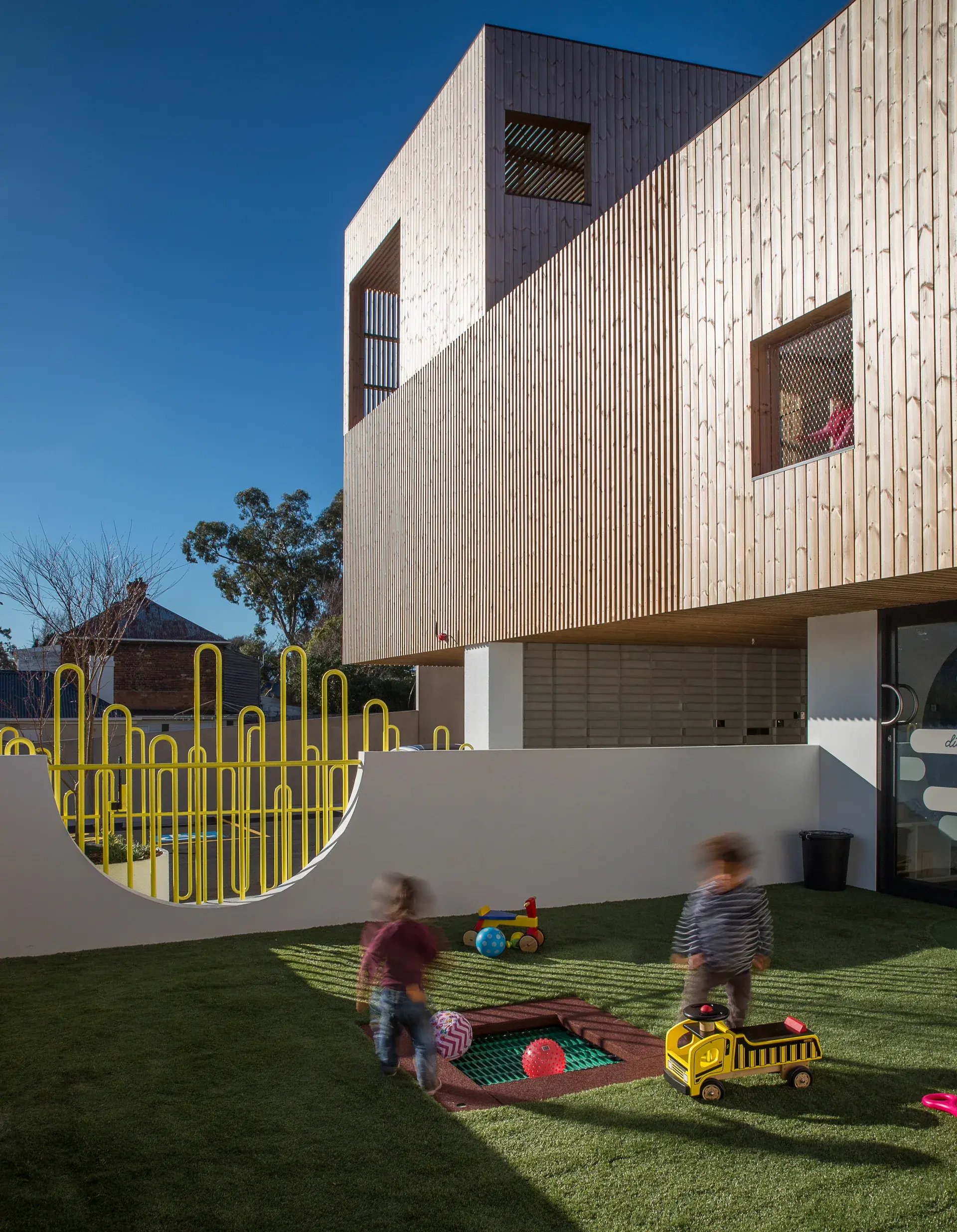 This outdoor play area features an artificial turf lawn and a unique yellow wavy fence set into a white wall. In the background, a modern building with vertical wood siding overlooks the space.