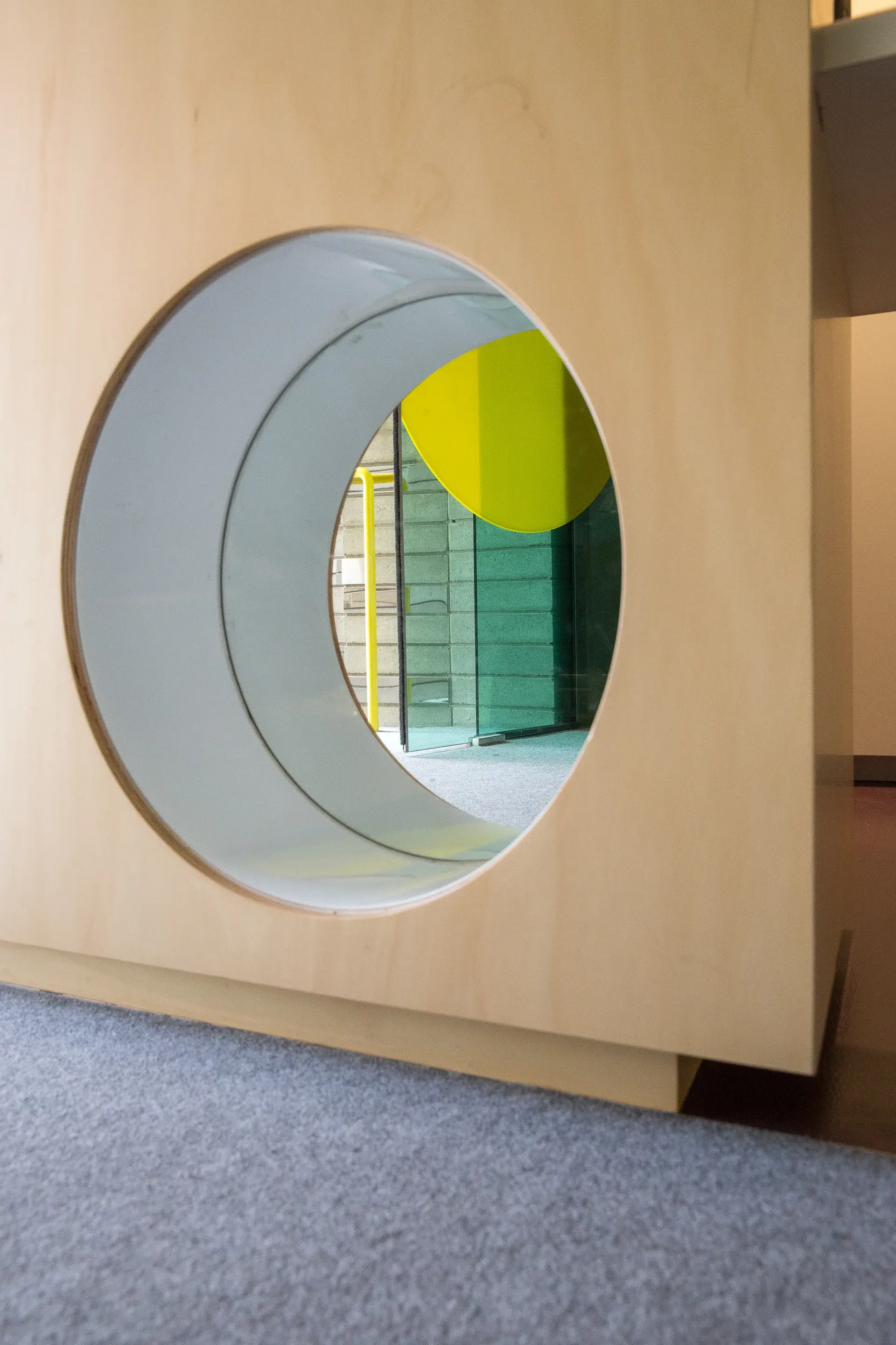 This close-up features a circular cutout in a light-wood structure, forming a small tunnel. A vibrant yellow element and glass partition are visible through the opening.