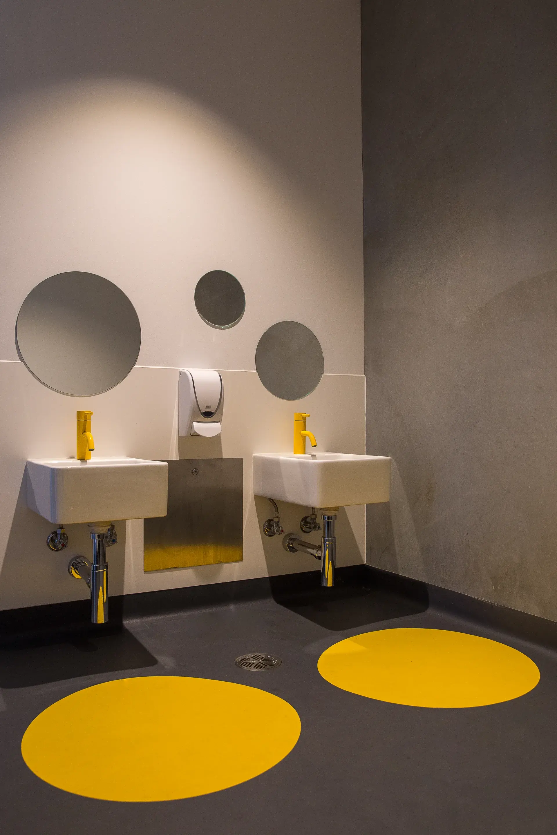 This bathroom features two white sinks with bright yellow faucets. Three circular mirrors hang above, and matching yellow circles are painted on the floor.