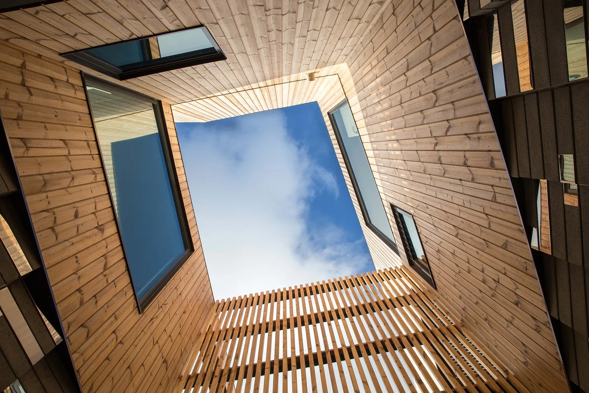 Looking up through the centre of the building, the view features wood-panelled walls and slatted screens surrounding a square opening to the sky. Several large windows are set into the facade, reflecting the clouds above.