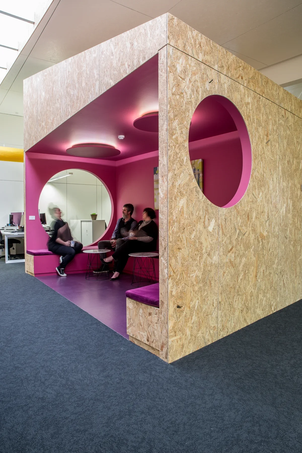 This structure is built from OSB (oriented strand board) with a vibrant pink interior and purple floors. It features large circular cutouts and cushioned bench seating where people are gathered.