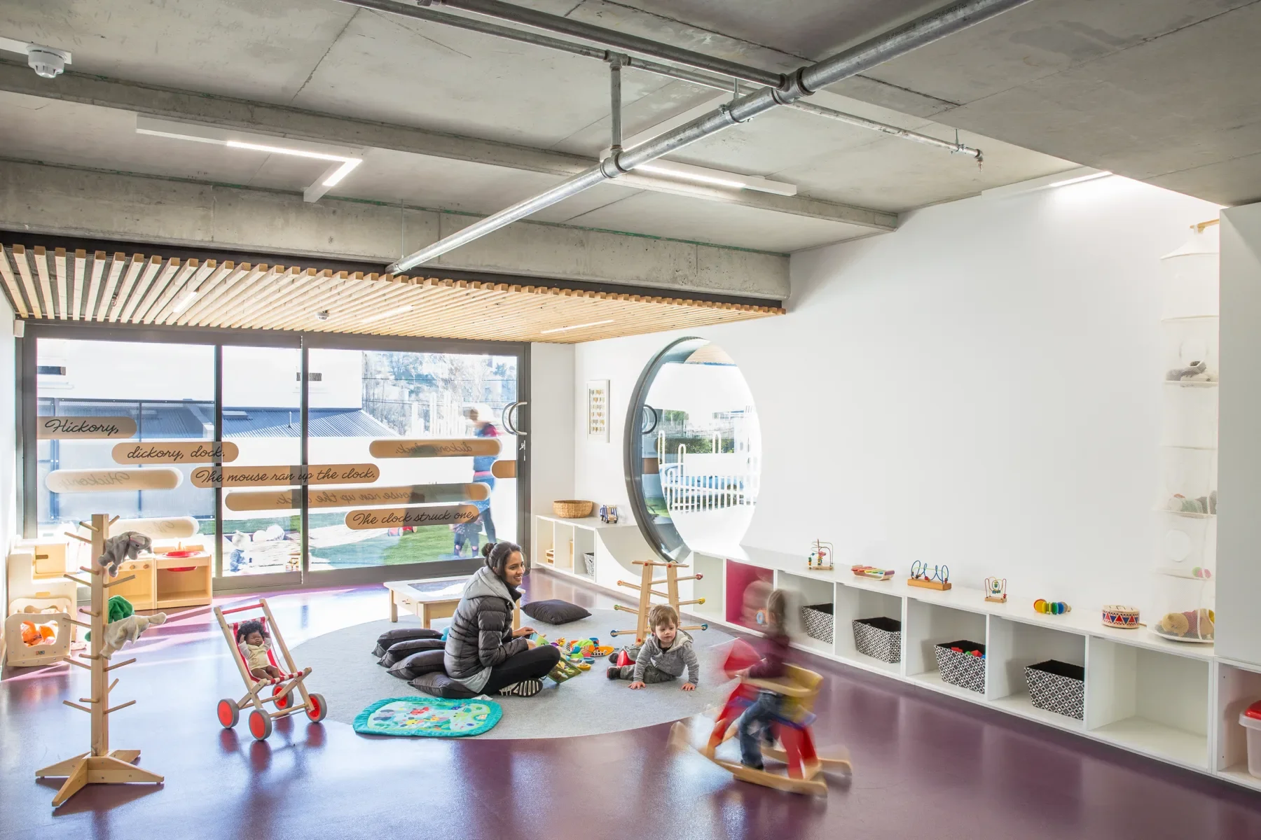 This bright playroom features a large circular window and glass doors with nursery rhyme text. A carer and children are playing on a grey rug surrounded by low white shelving and various wooden toys.