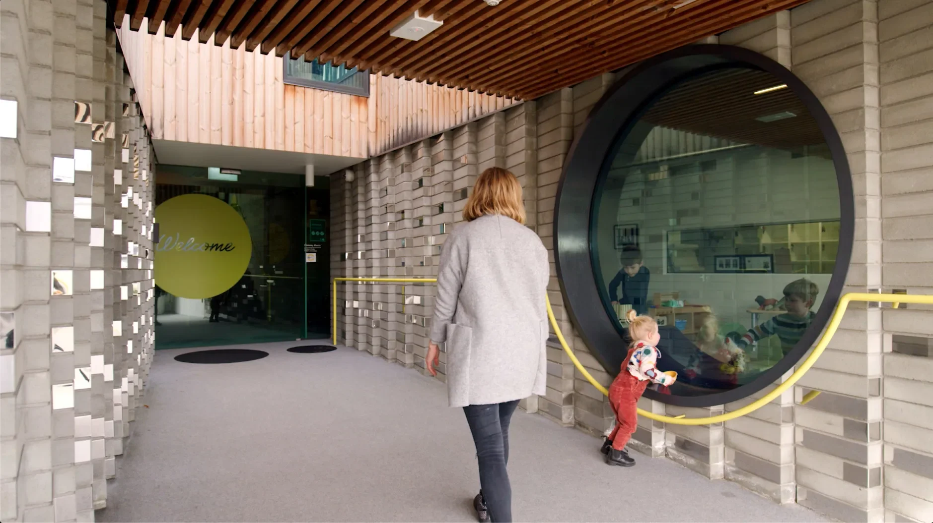This hallway features grey brick walls with reflective inserts and a large circular window looking into a playroom. A yellow railing follows the curve of the window, and a woman walks toward a glass door marked with a large yellow "Welcome" circle.