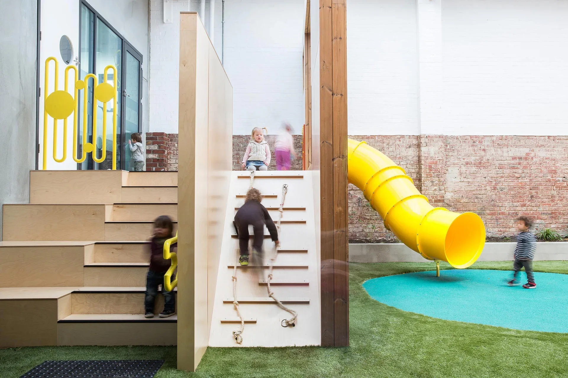 This outdoor play area features a yellow slide and a wooden climbing ramp with ropes. Children are playing on the equipment and a set of wide wooden steps, which are accented by a vibrant yellow railing.