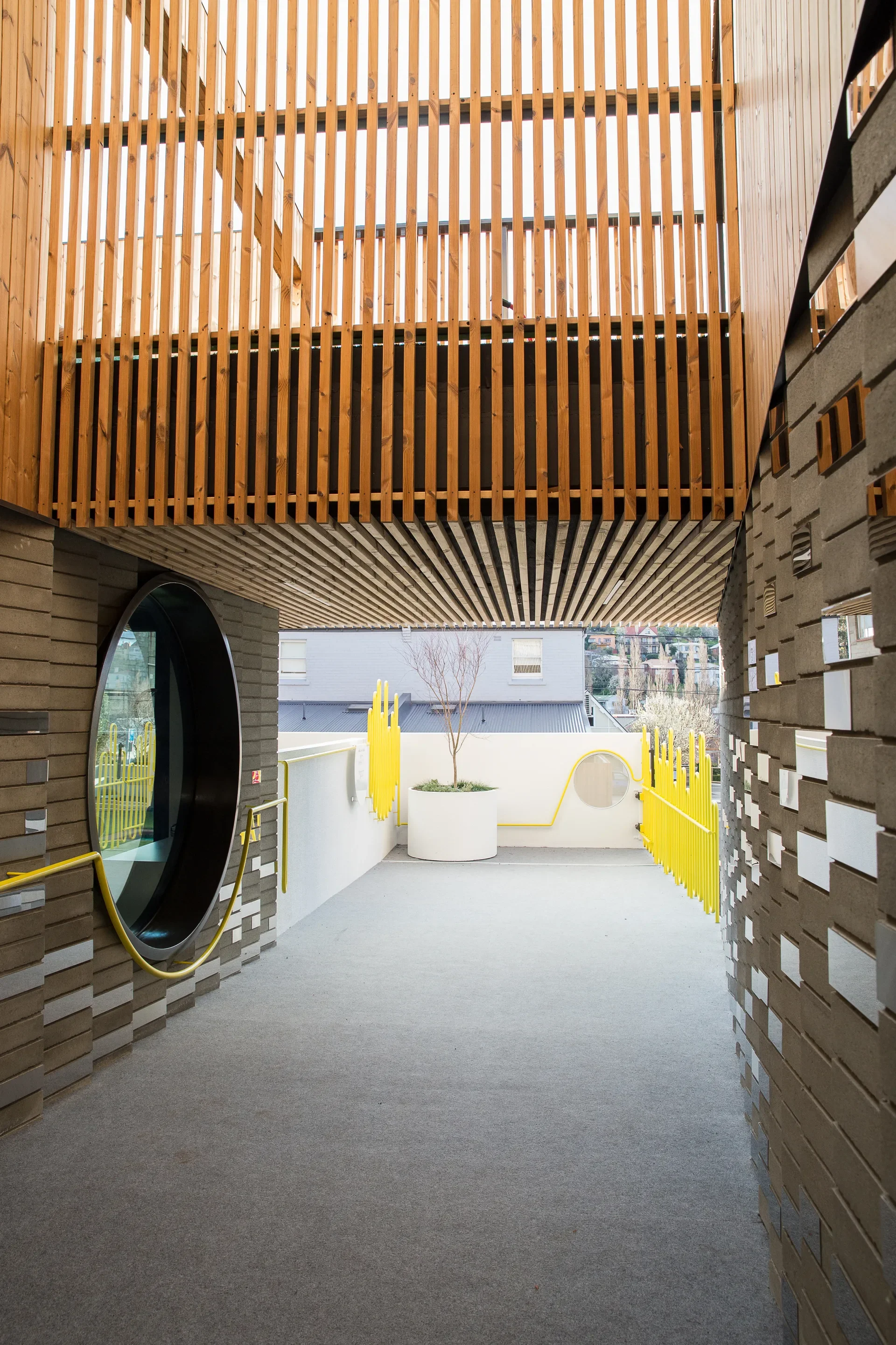 This corridor features grey brick walls with reflective inserts, a slatted wood ceiling, and a large circular window. A yellow railing leads toward a white planter and a matching vibrant yellow fence at the walkway's end.