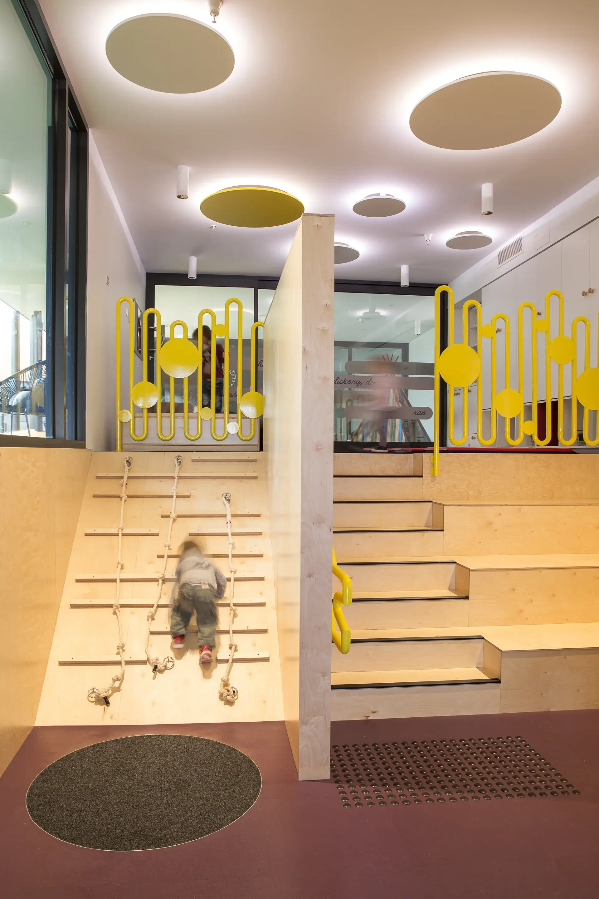 This indoor play area features a light-wood climbing ramp with ropes and a set of wide wooden steps. Vibrant yellow railings with a wavy design border the top level, while large circular light fixtures glow on the ceiling above.